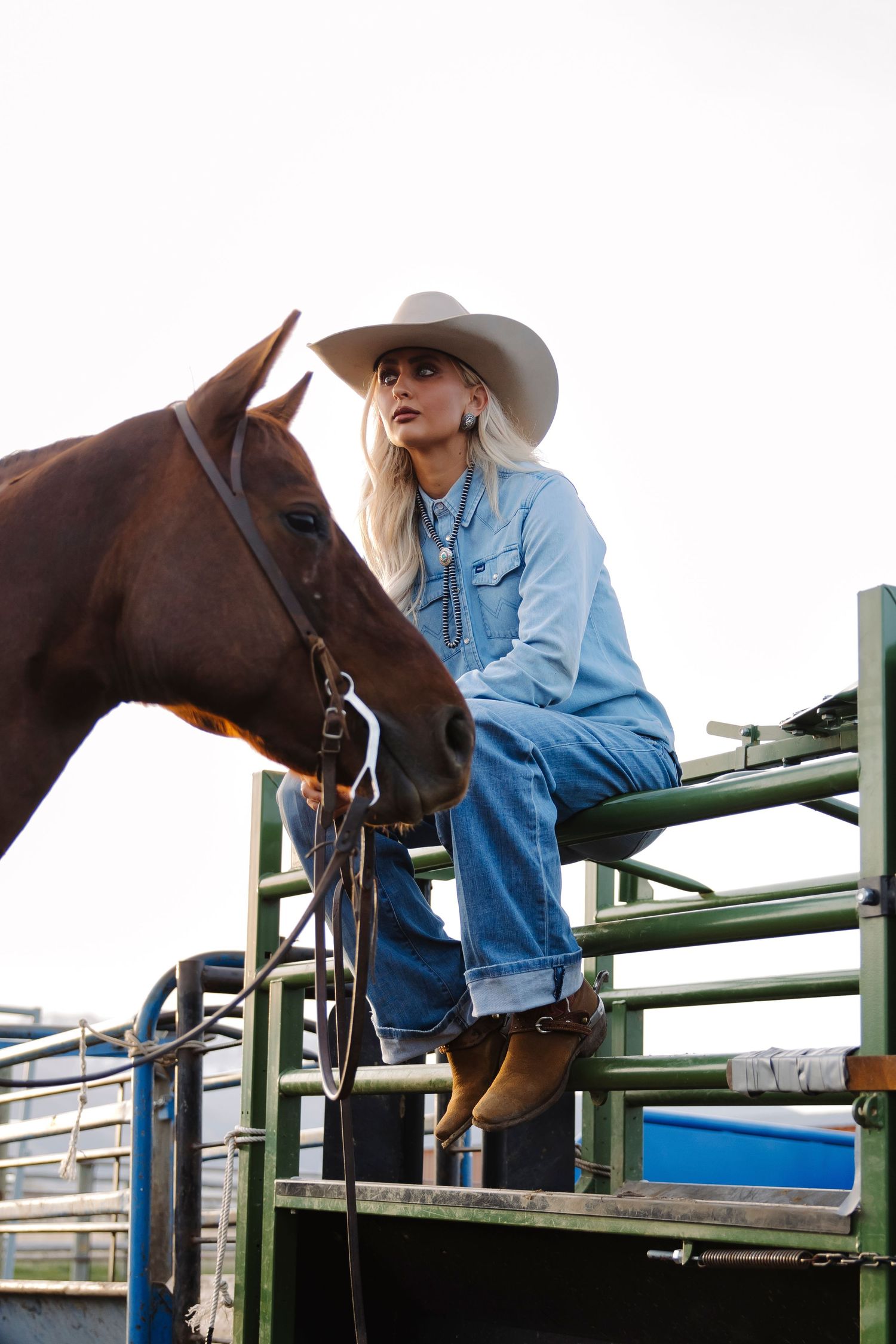Blonde woman in denim outfit and white cowboy hat sitting on ranch fence beside a brown horse.