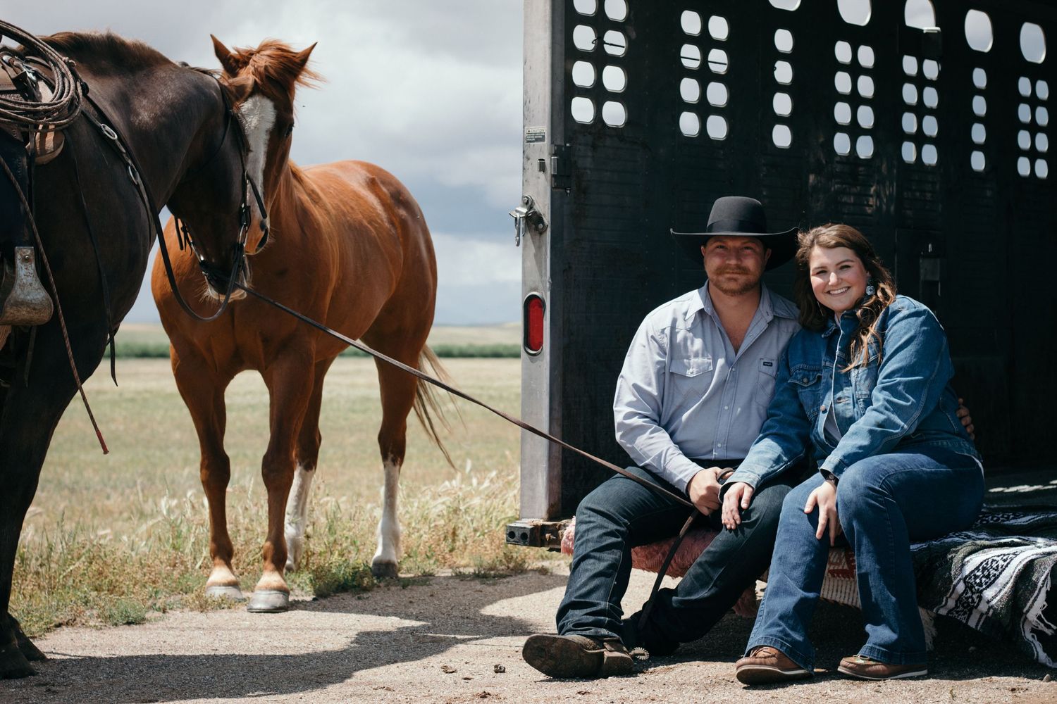 A couple sitting in a stock trailer with horses in the background.