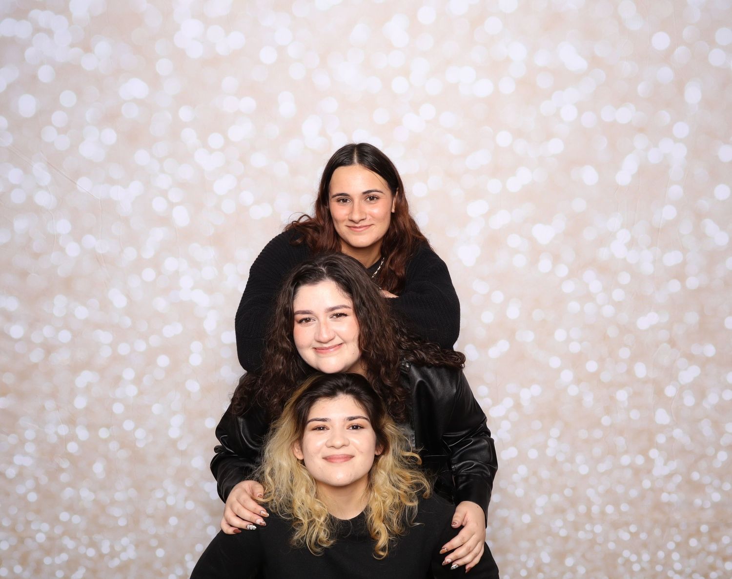 Three young women posing together in front of a bokeh light backdrop, wearing black outfits.