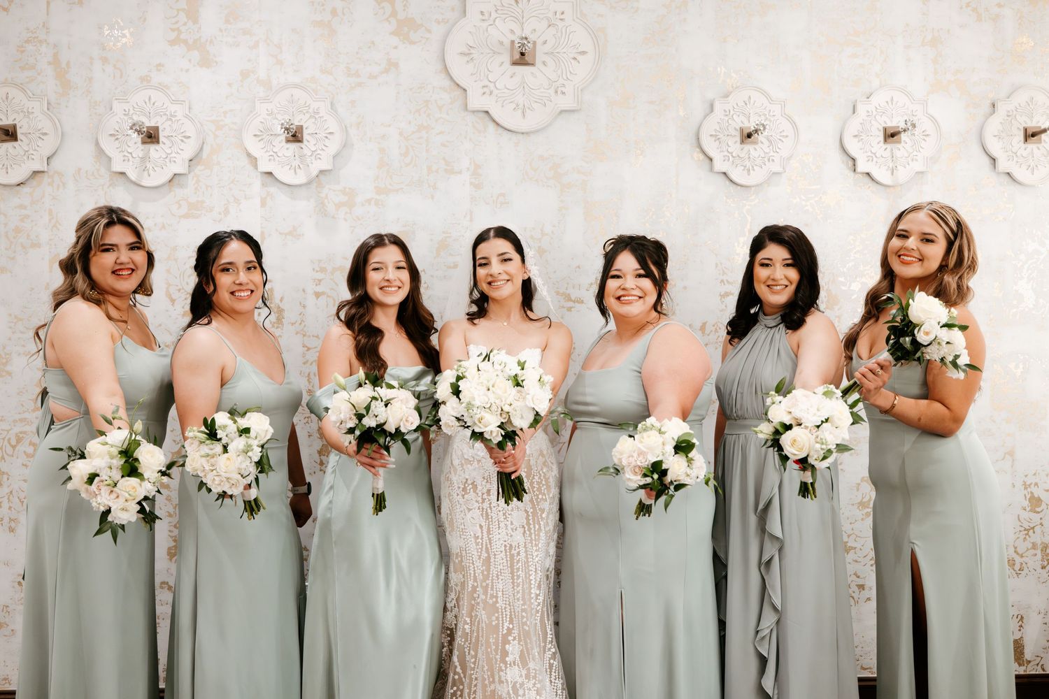 Bridal party in sage green dresses poses with bouquets against floral wall.