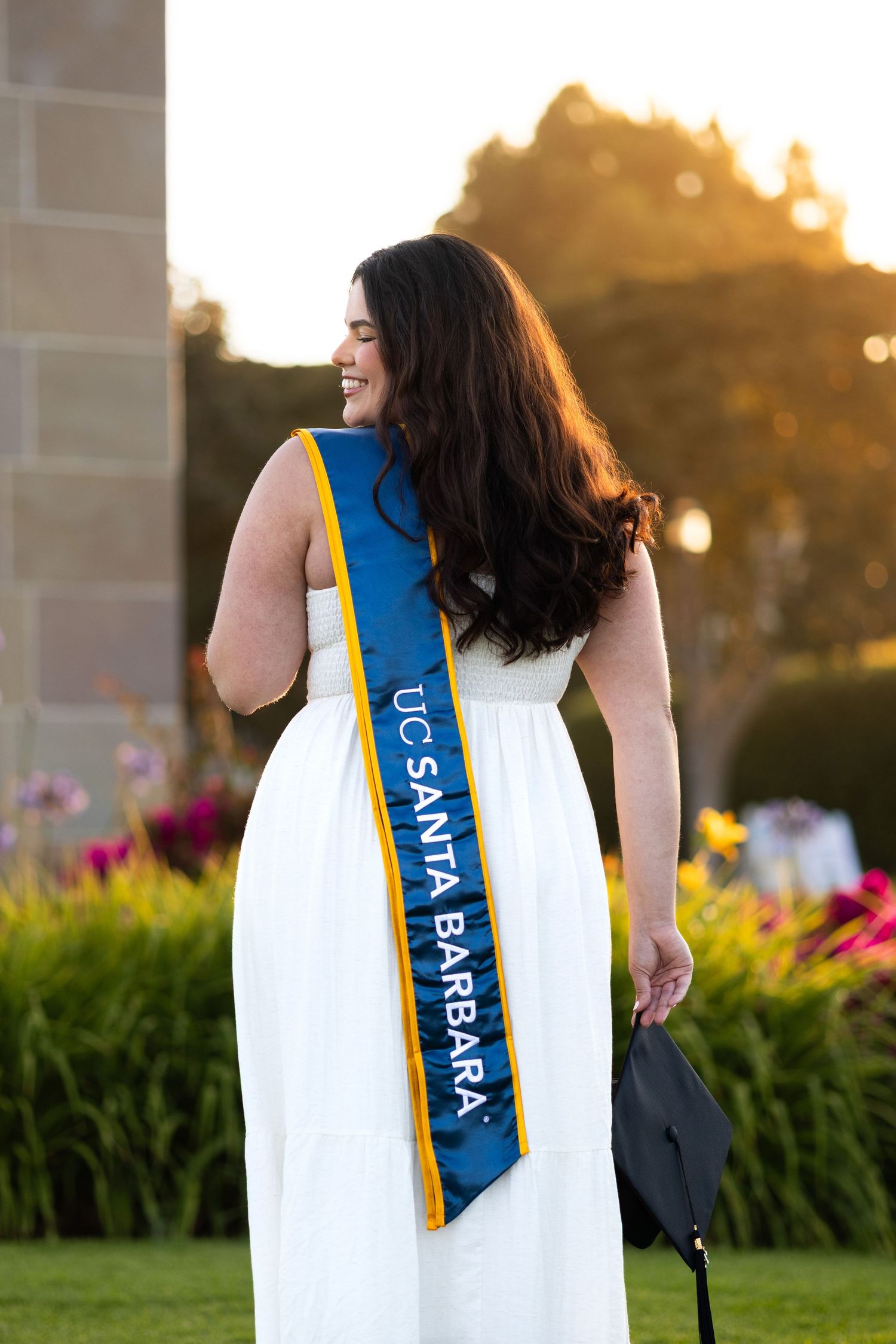 Graduate in white dress with blue and gold sash photographed against garden backdrop at sunset.
