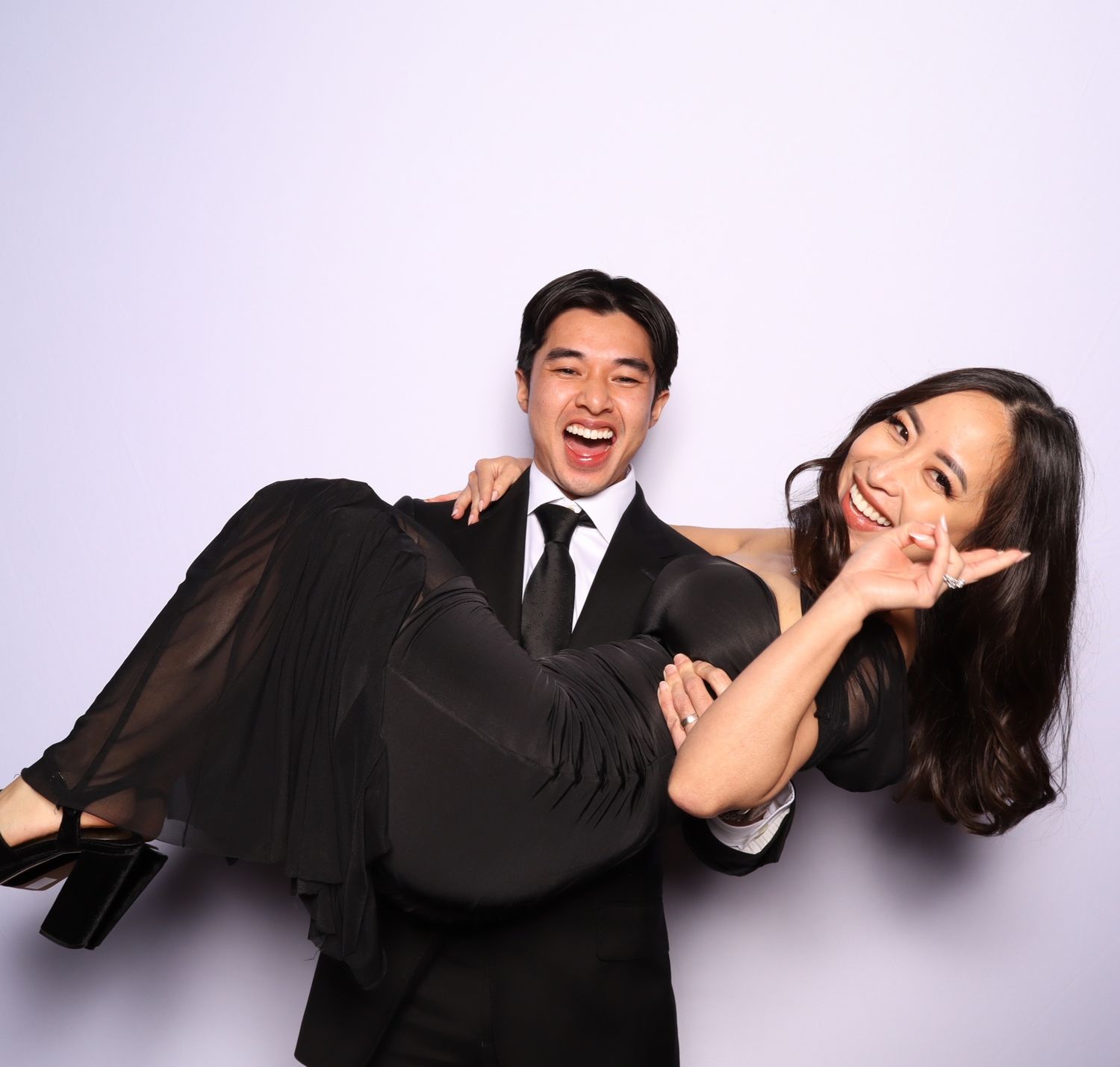 A smiling man in a black suit carries a laughing woman in a black dress at a formal event photo booth.