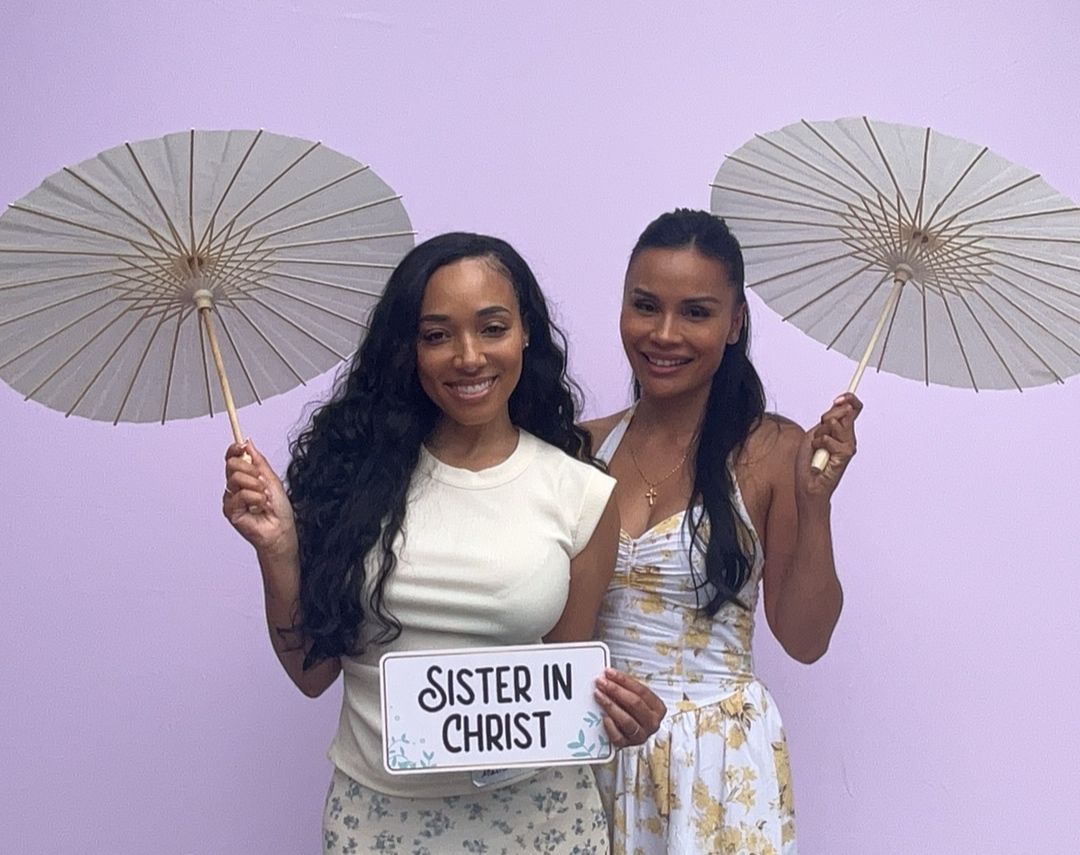Two people in white outfits pose with decorative white parasols during what appears to be a wedding celebration.