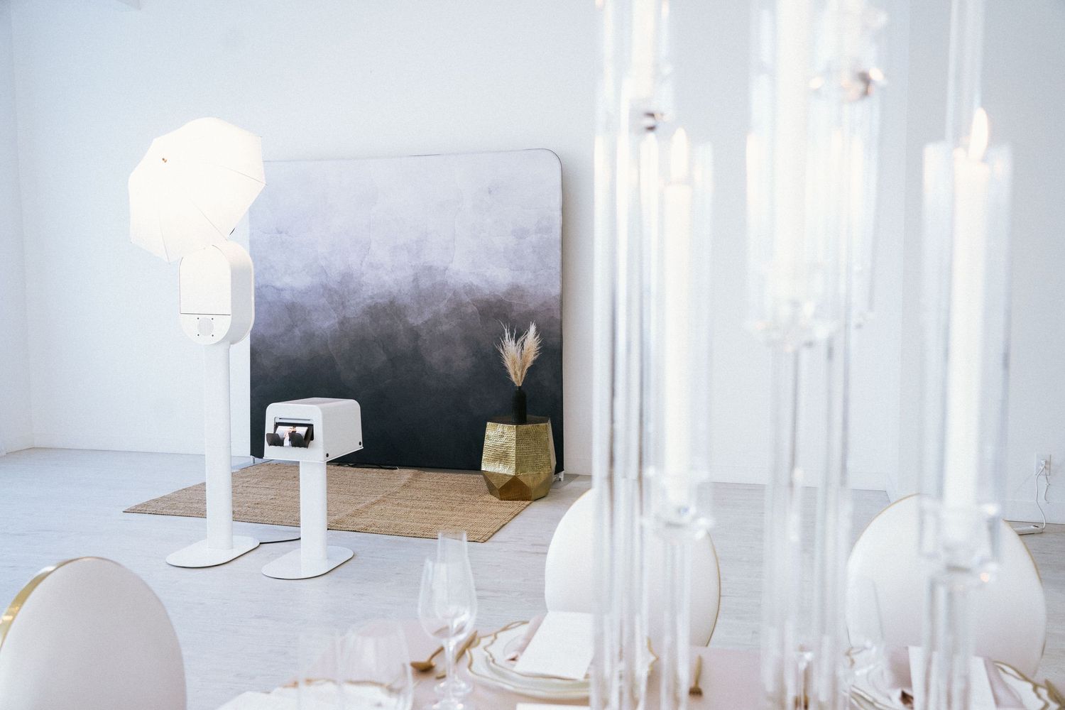 Elegant white event table setting with glass candleholders, white chairs, and an ombre art backdrop with gold decor.