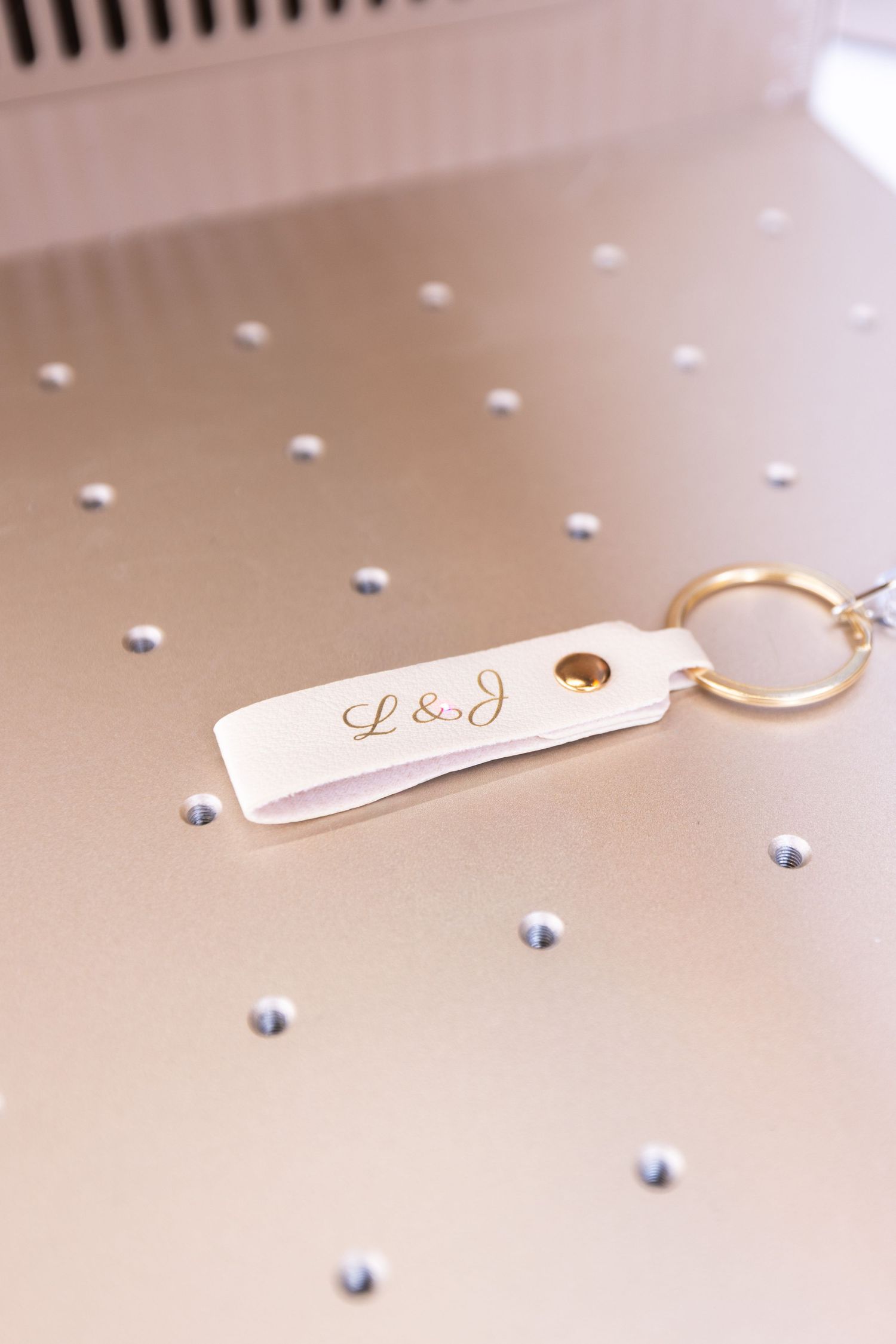White leather keychain engraved with L&J initials and gold ring on decorative surface with rhinestone accents.