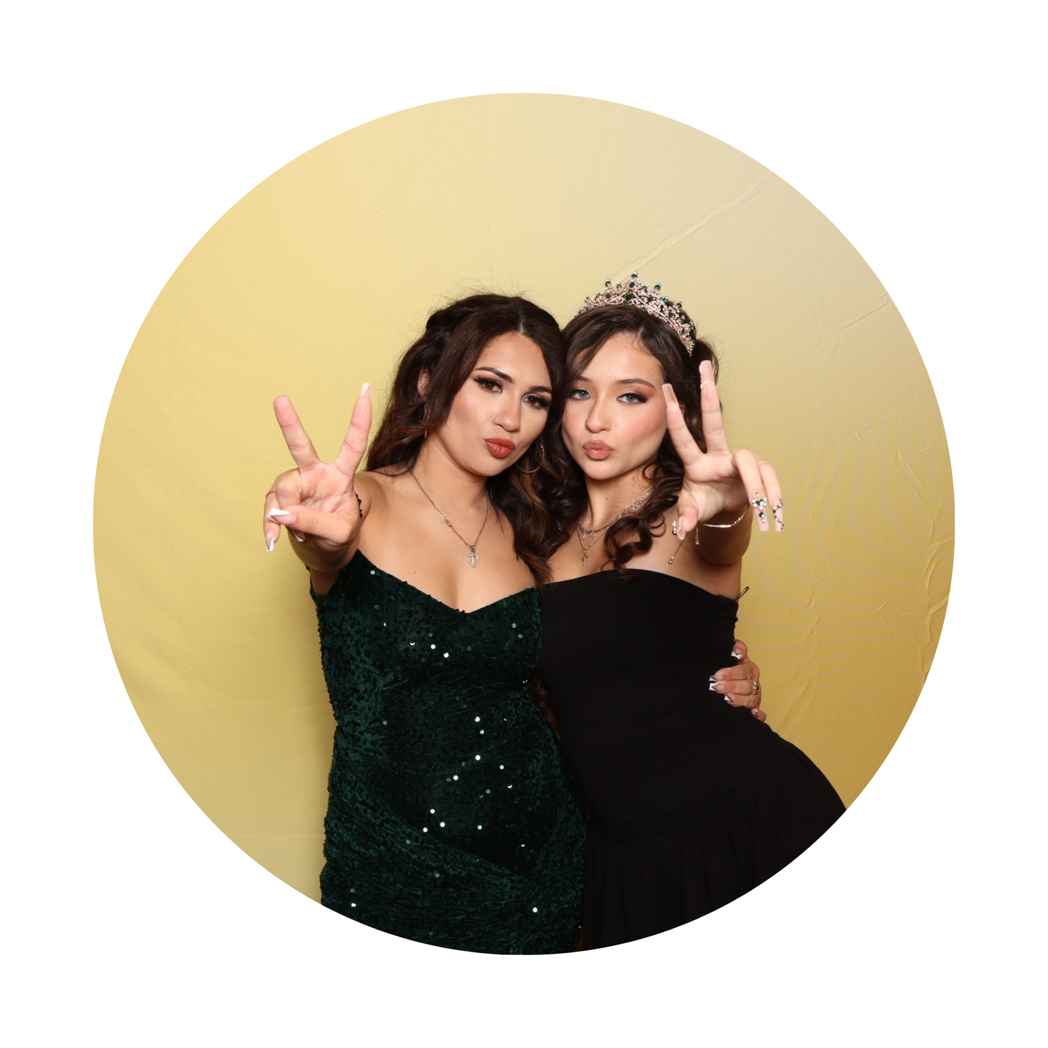 Two young women in formal attire pose with peace signs, one wearing a crown and black dress, the other in a green sequin gown.