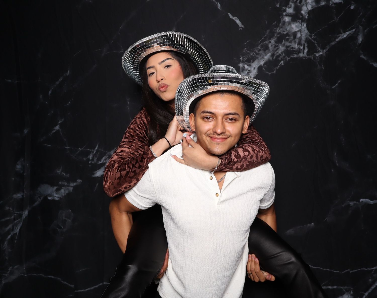 Two friends posing at a party, one giving the other a piggyback ride, both wearing matching silver bucket hats.