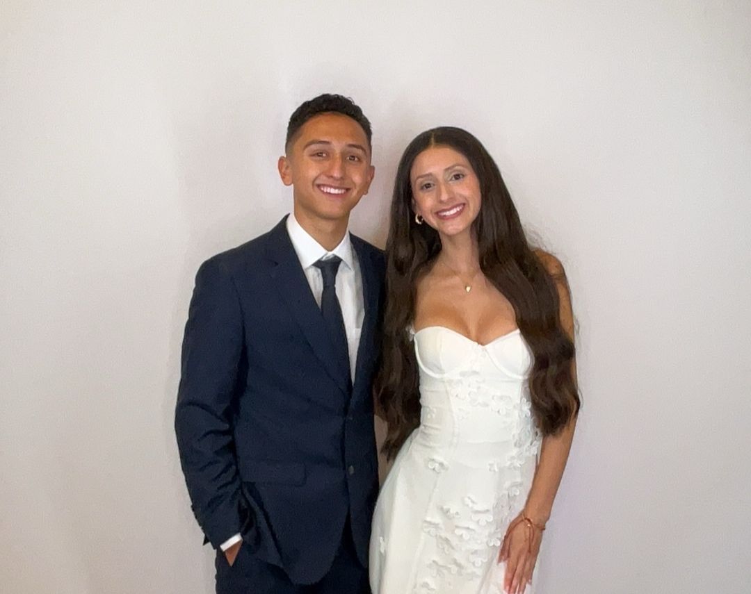 A smiling couple poses together, man in navy suit and woman in white floral strapless dress, against a white background.