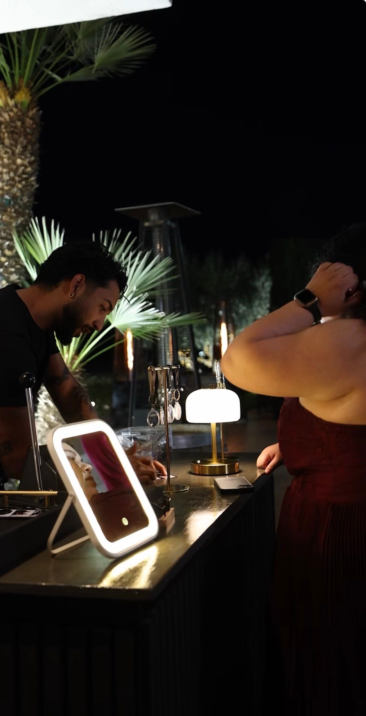 A makeup artist applies cosmetics at an illuminated vanity mirror station outdoors at night with palm trees in background.