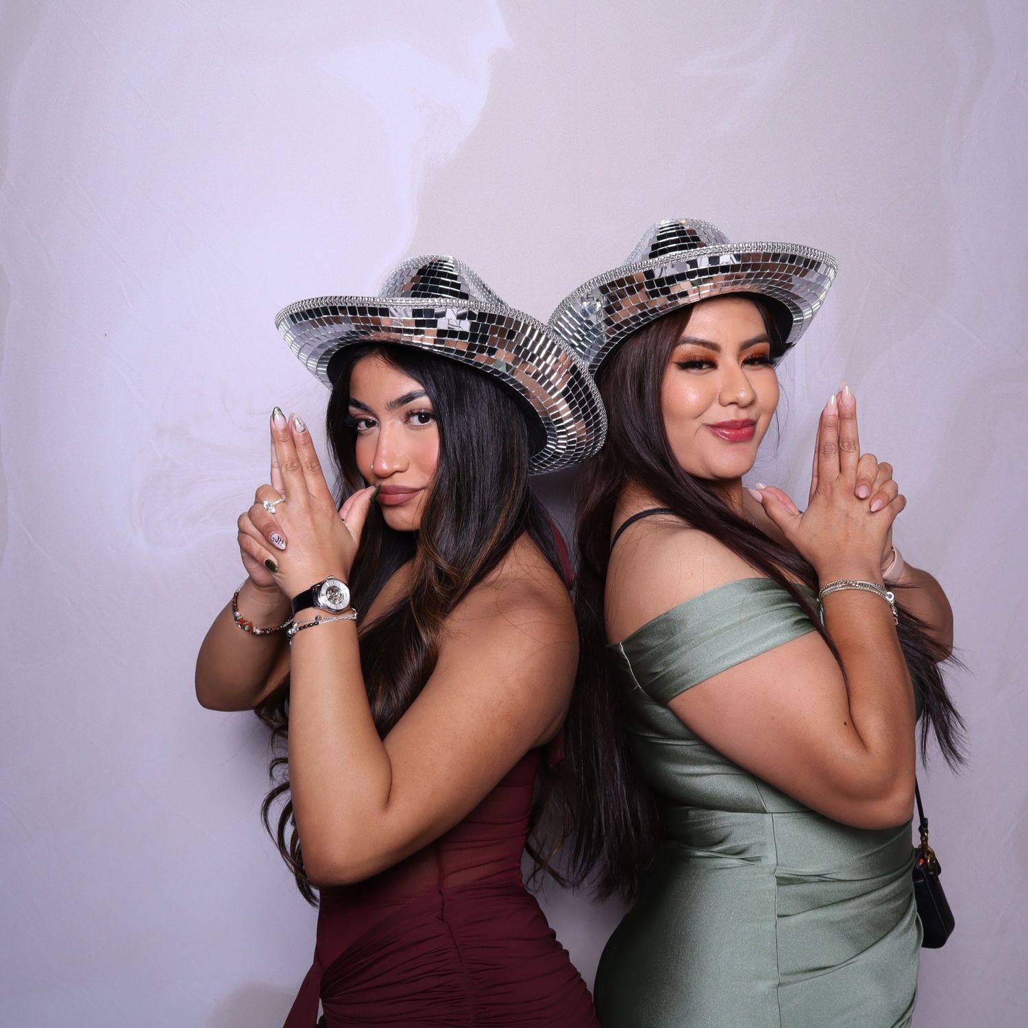 Two women in disco ball hats pose back-to-back in elegant dresses at a festive photo booth event.