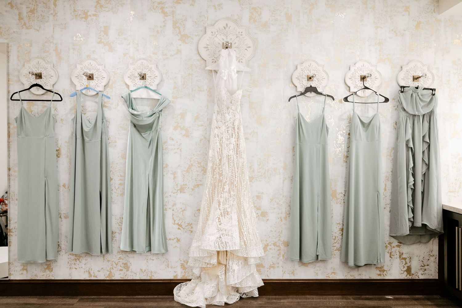 Elegant sage green bridesmaid dresses and a white wedding gown hanging on display against a textured wall.