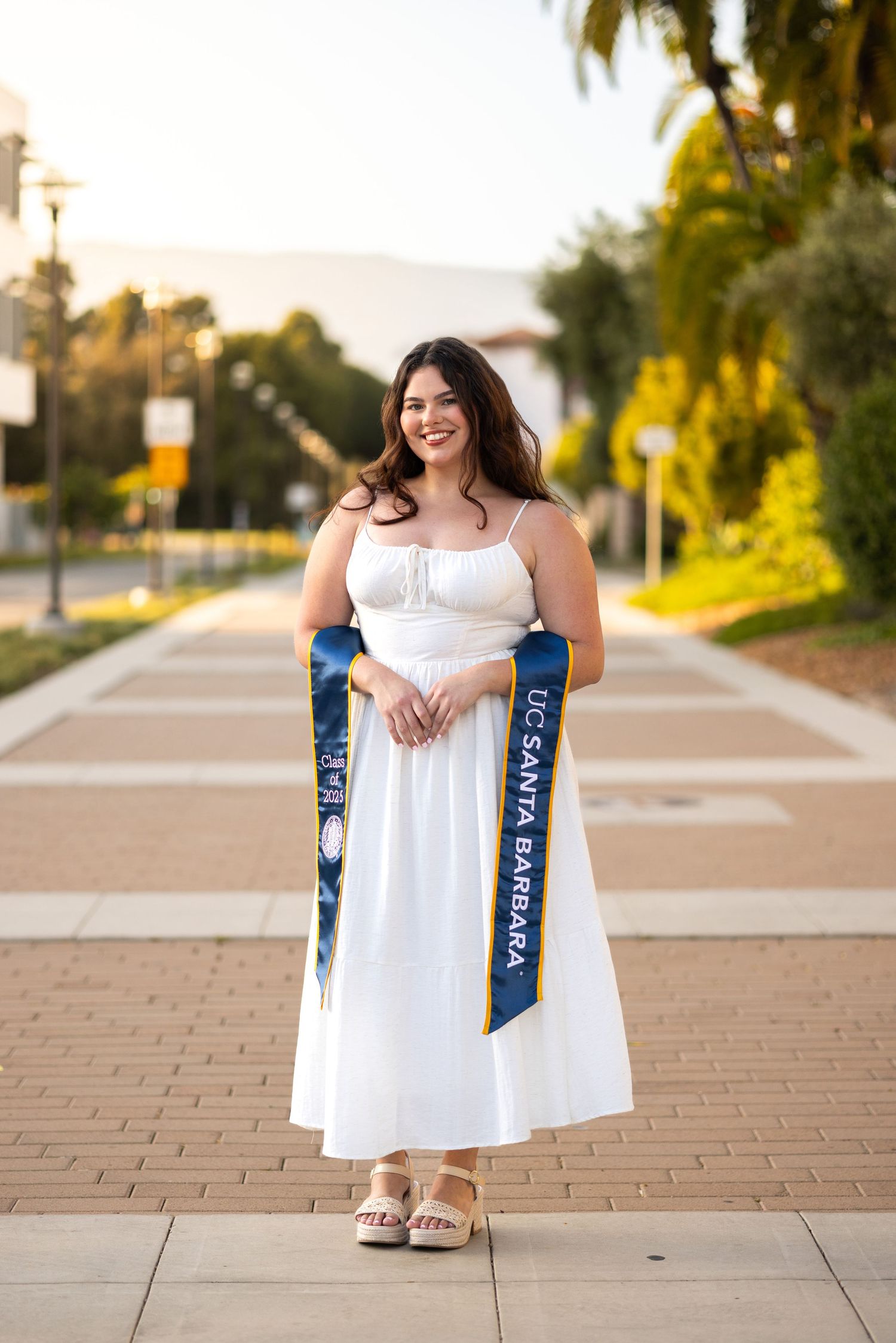 Graduate in white sundress with blue sash poses on stone pathway during golden hour.