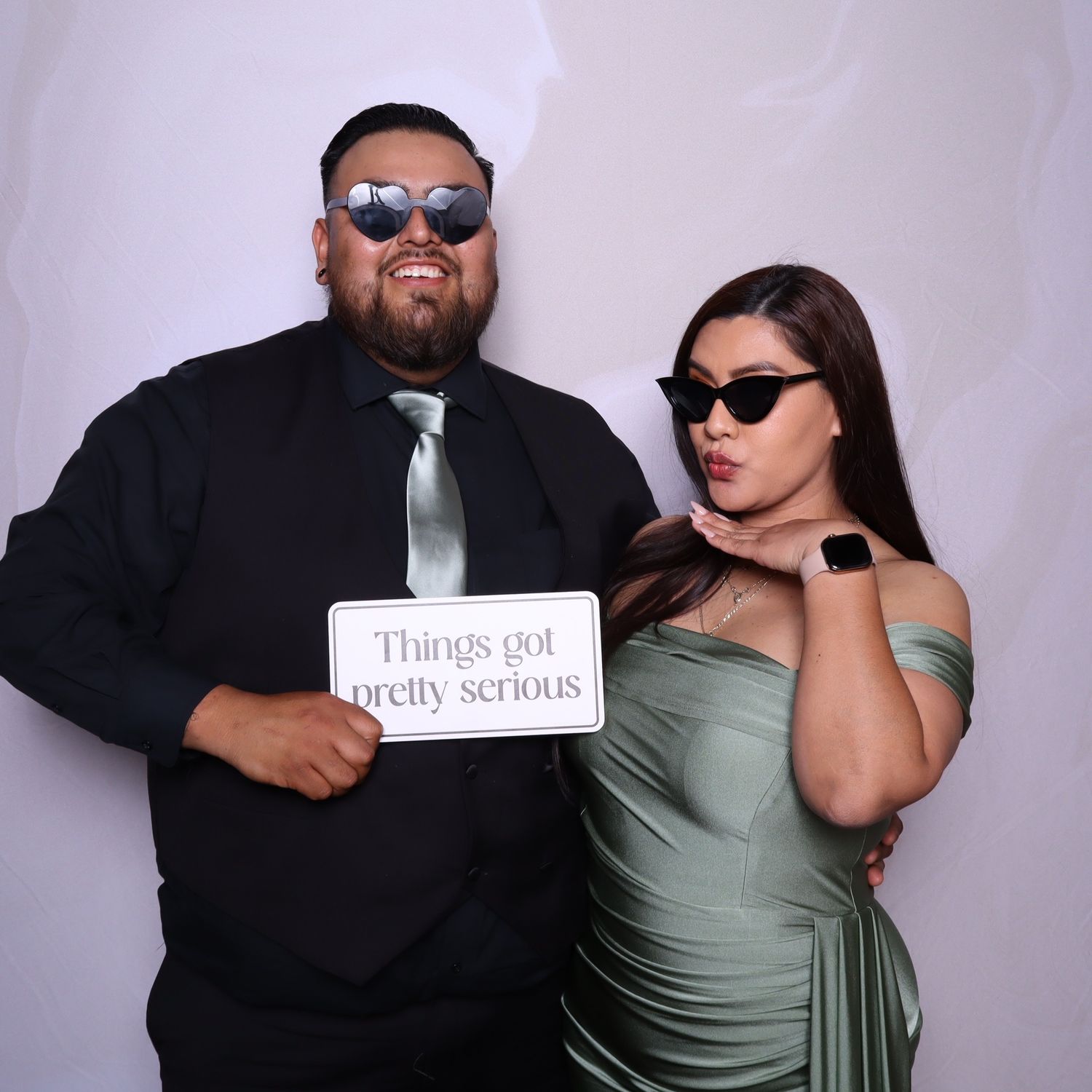 Couple posing in photo booth, man in black suit with tie, woman in sage green dress, both wearing sunglasses, holding funny sign.