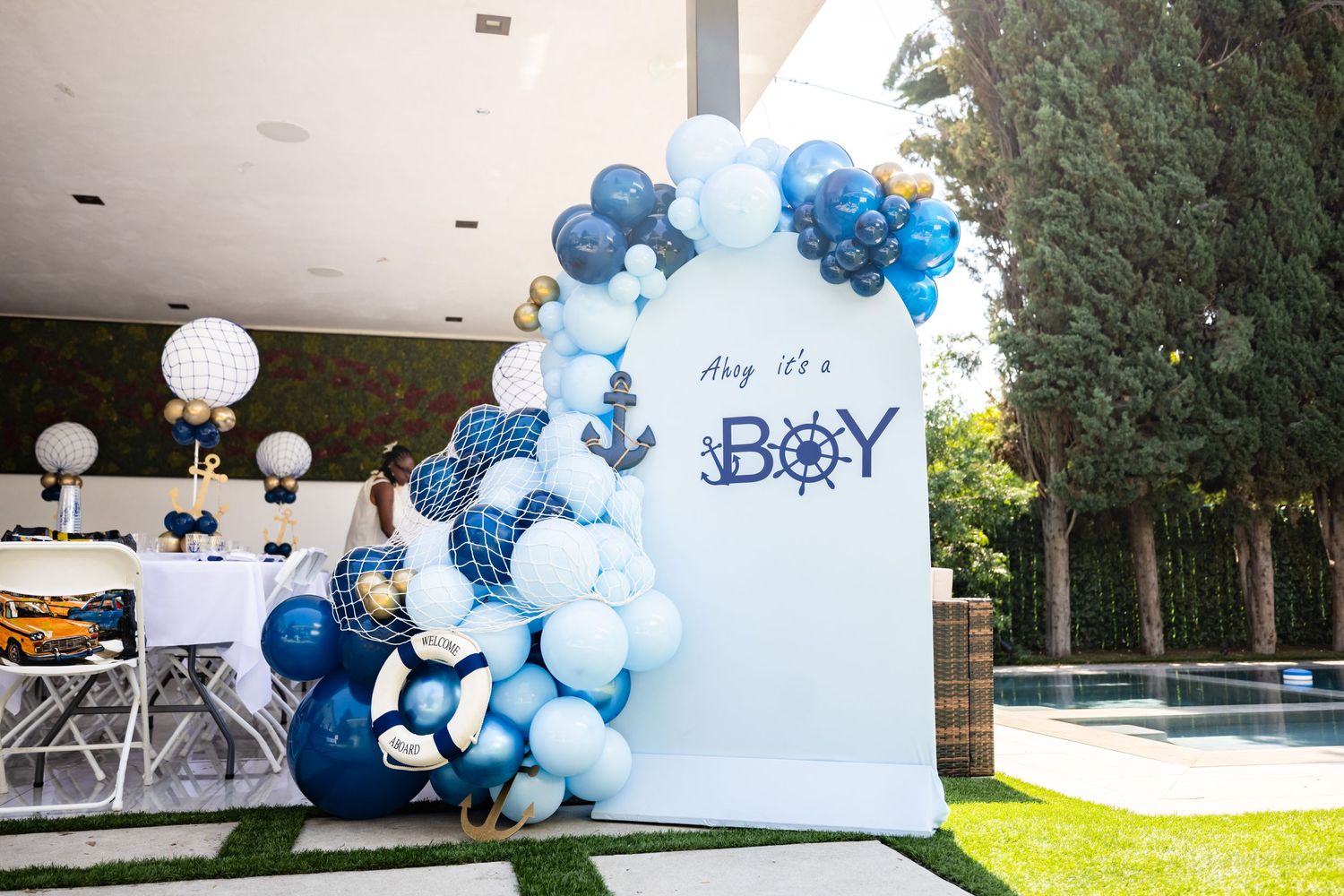 A nautical themed baby shower backdrop with blue and white balloon arch and anchor decorations.