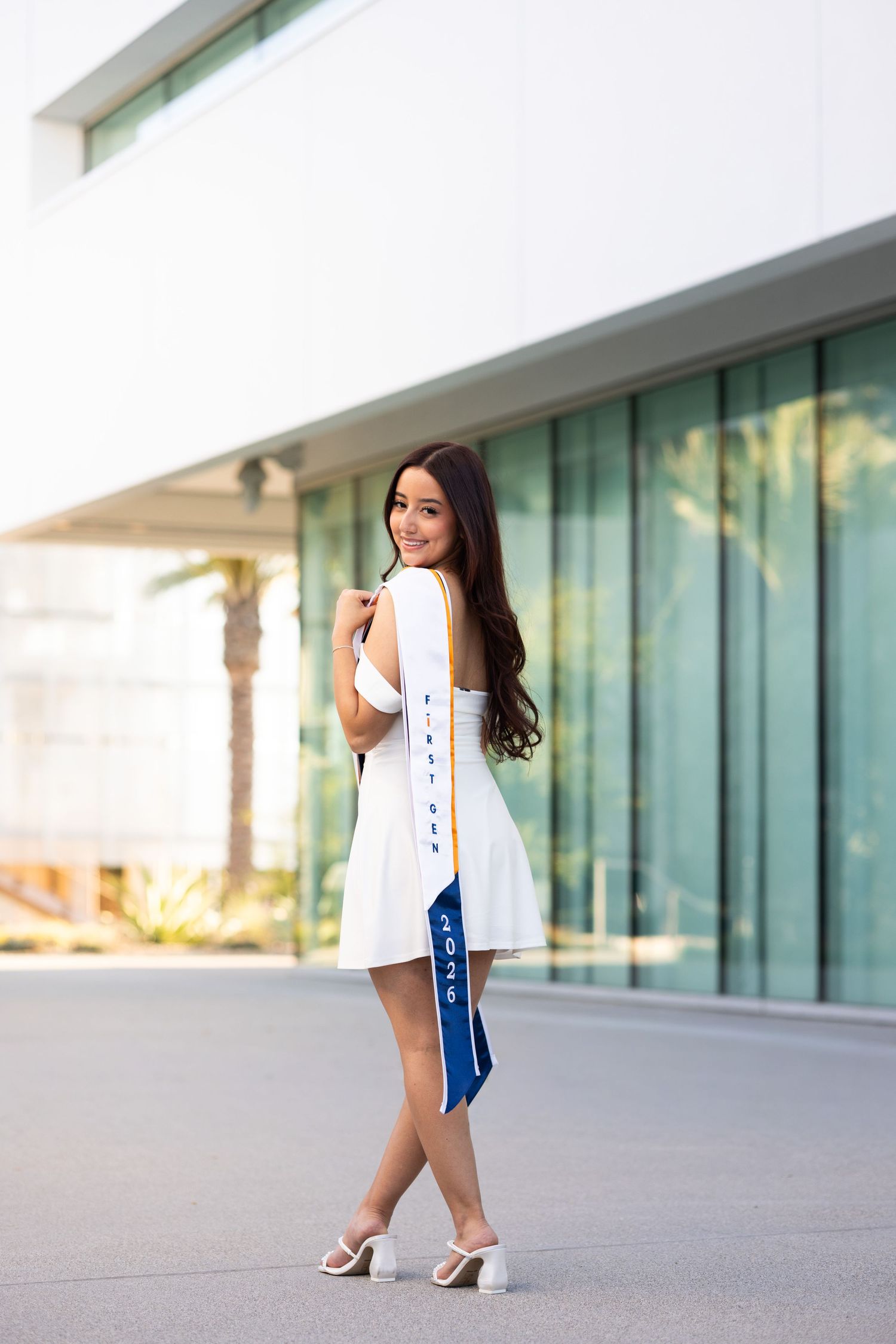 Young woman in white dress holding blue graduation stole, posing outdoors near modern building for senior portrait.