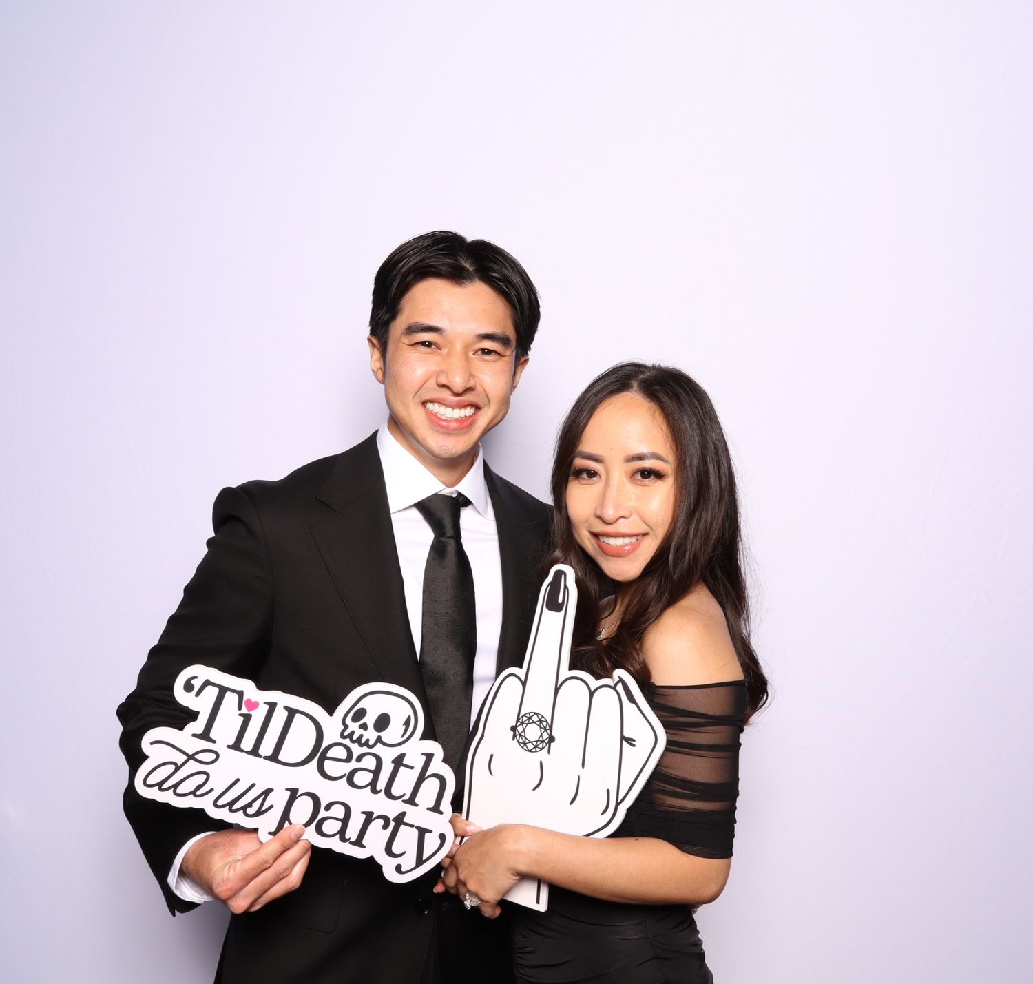 Couple posing in photo booth holding fun party props with 'Til Death Do Us Party' sign at wedding reception.