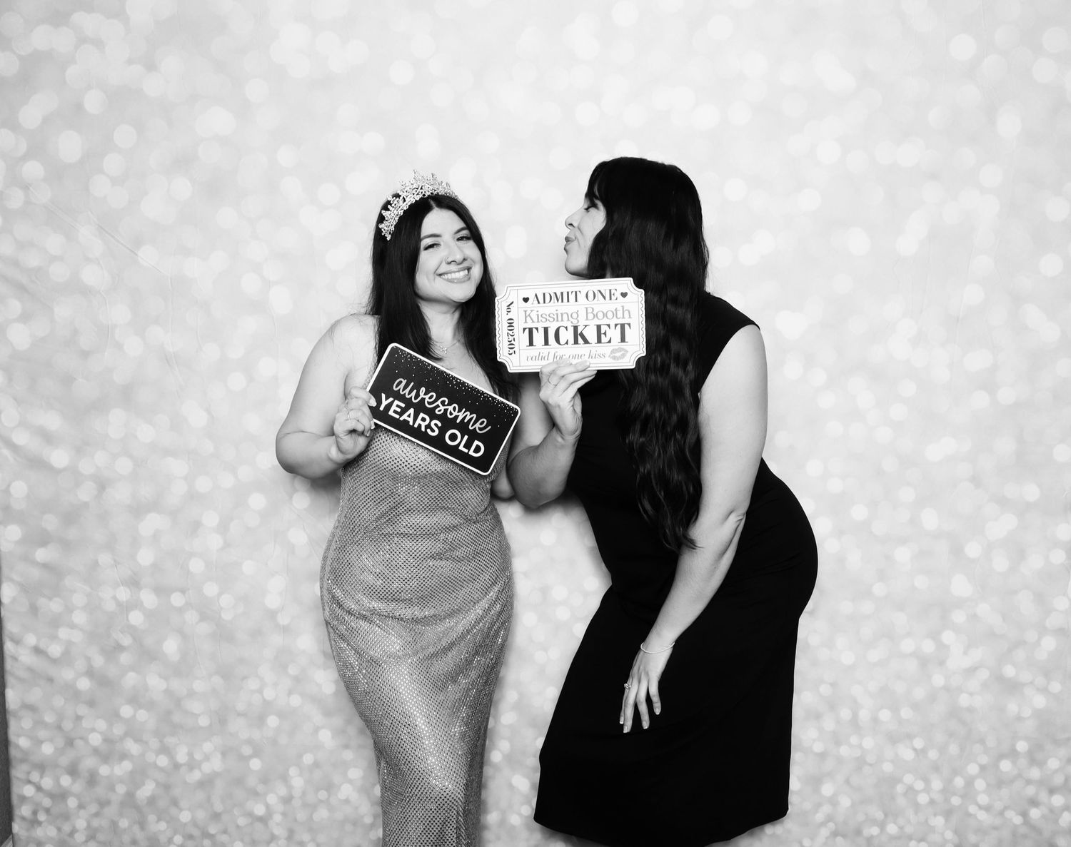Two women posing in a photo booth, one in sequin dress with birthday sign, other in black dress holding ticket sign.
