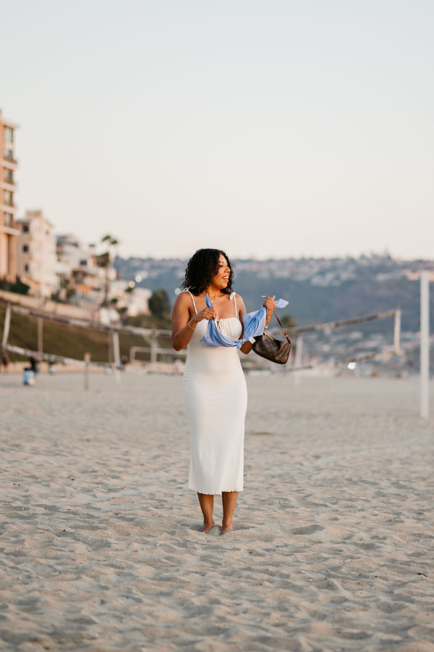 A woman in a white dress poses elegantly on a sandy beach at sunset with coastal buildings in the background.