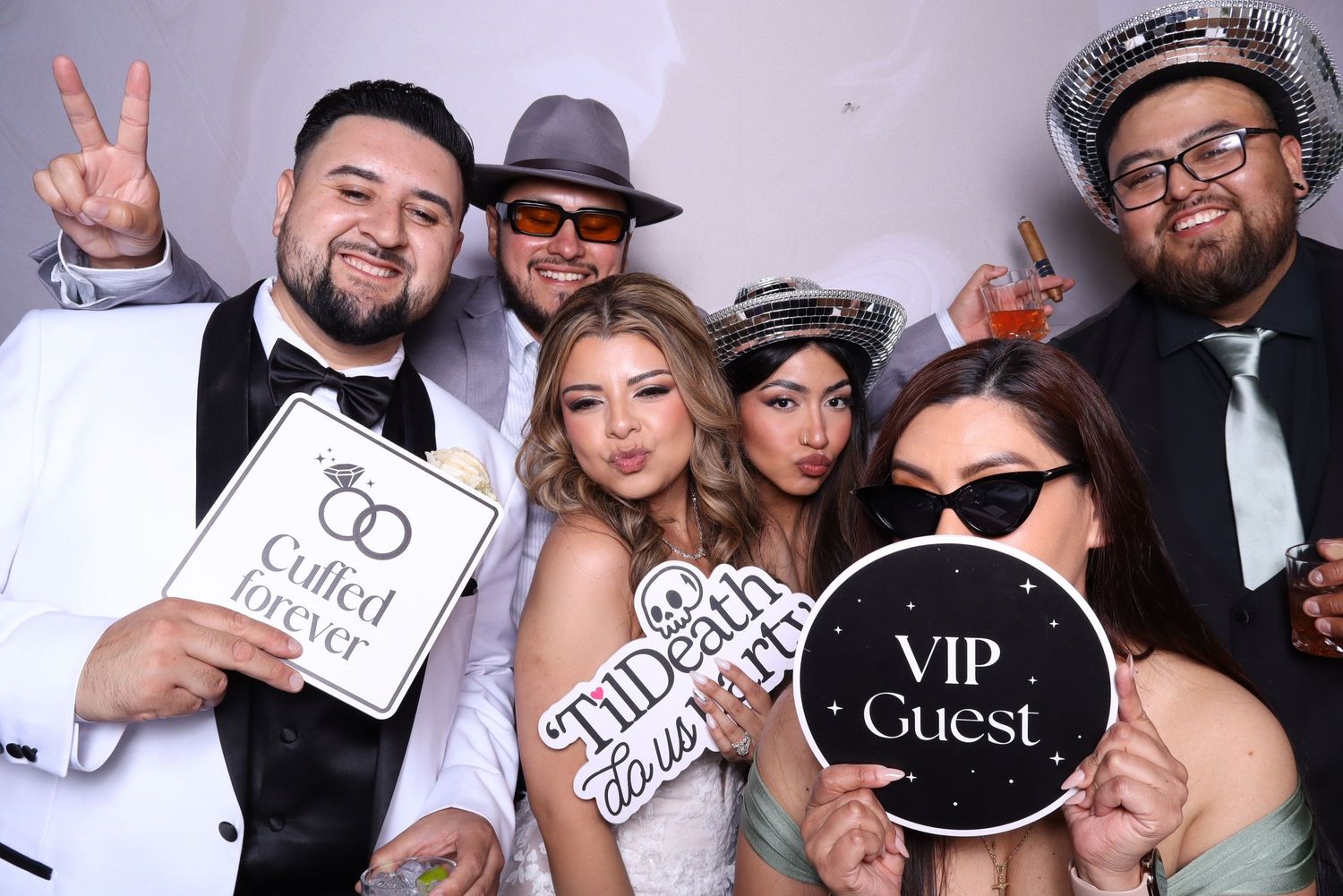 Group of six friends posing at a photo booth with fun signs including VIP Guest and Til Death at a wedding celebration.