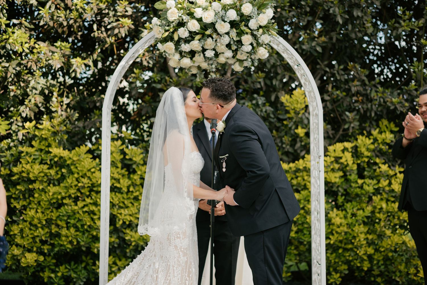 A romantic wedding ceremony moment under a white floral arch in a garden setting.