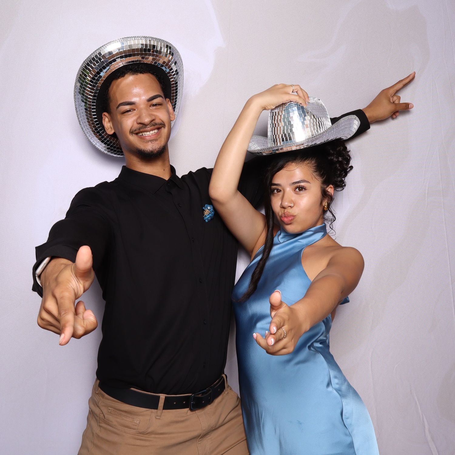 Two people pose playfully wearing disco ball hats, one in black outfit and one in blue dress, pointing at camera.
