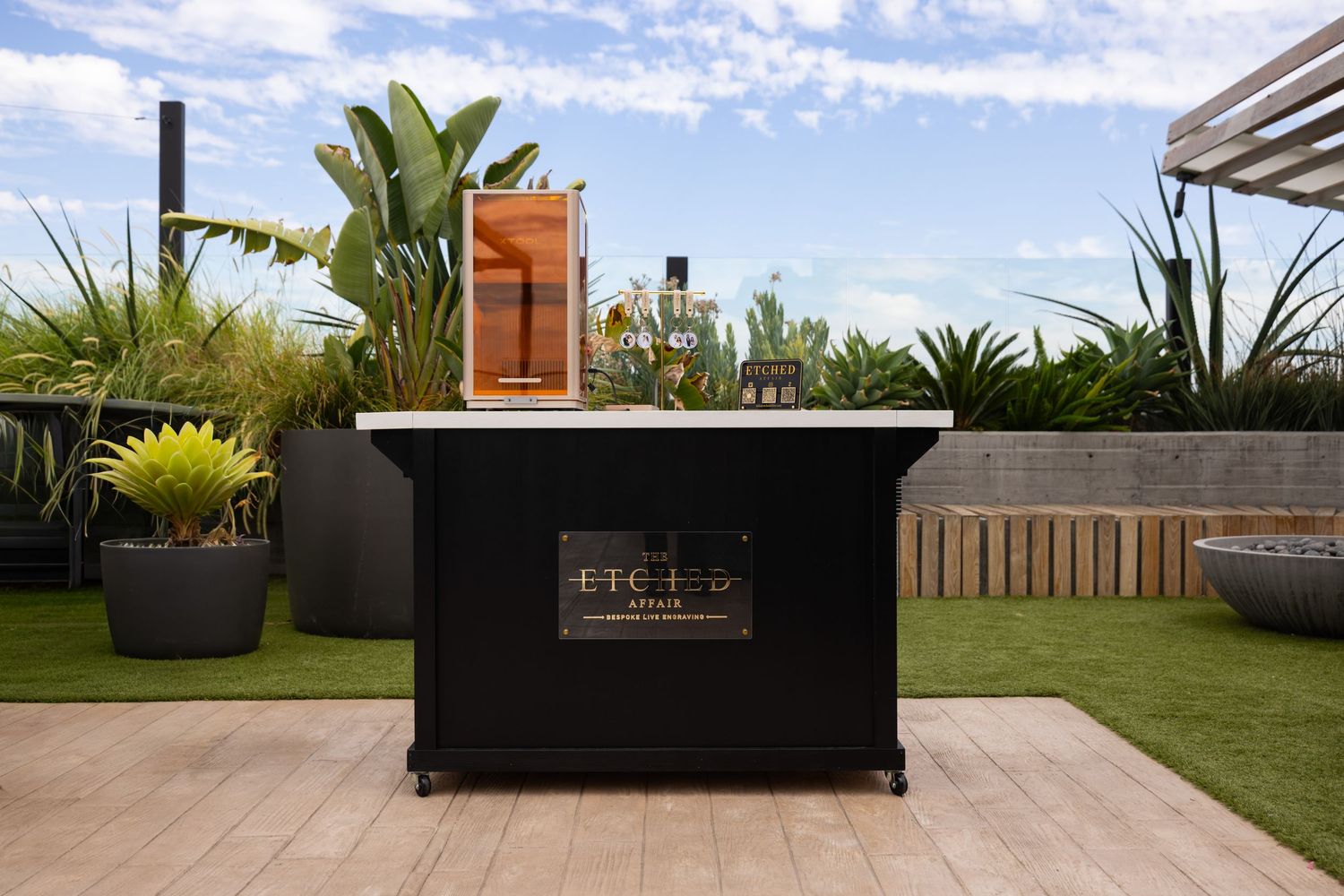 Etched luxury outdoor bar cart with marble top displayed on a wooden deck surrounded by lush greenery.