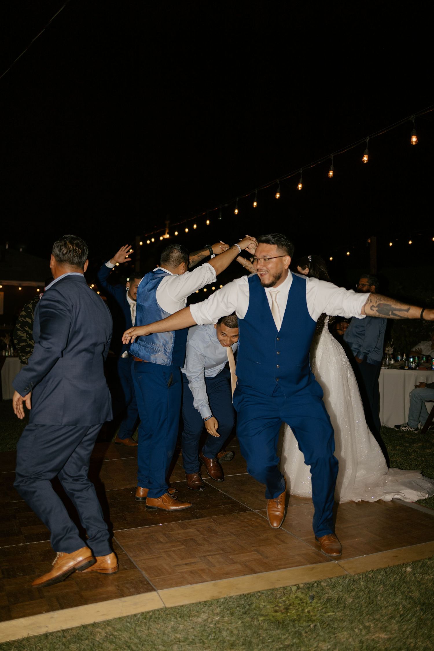 Wedding guests dance energetically on an outdoor dance floor under string lights at night.