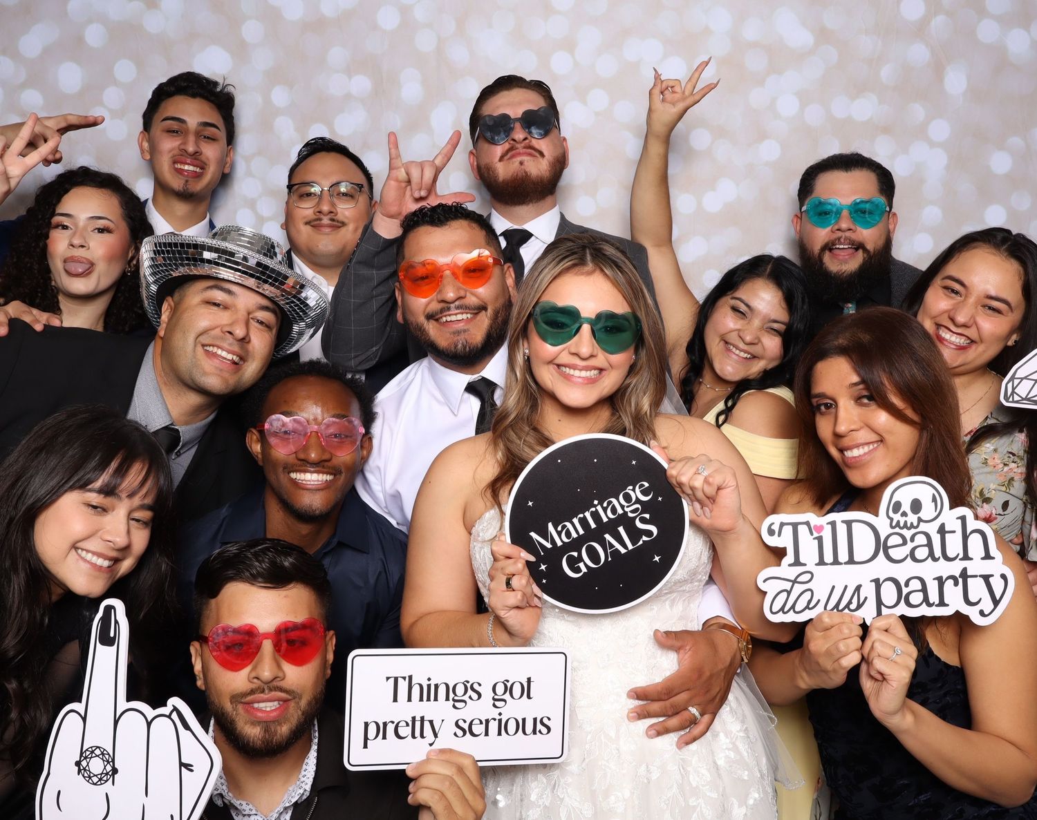 A festive group photo booth shot with people wearing colorful sunglasses and holding fun party props against a sparkly backdrop.