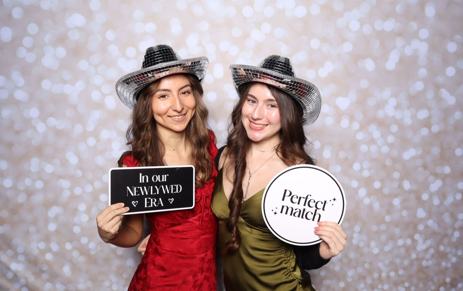 Two women in party hats pose at a photo booth holding signs reading Newlywed Era and Perfect Match.