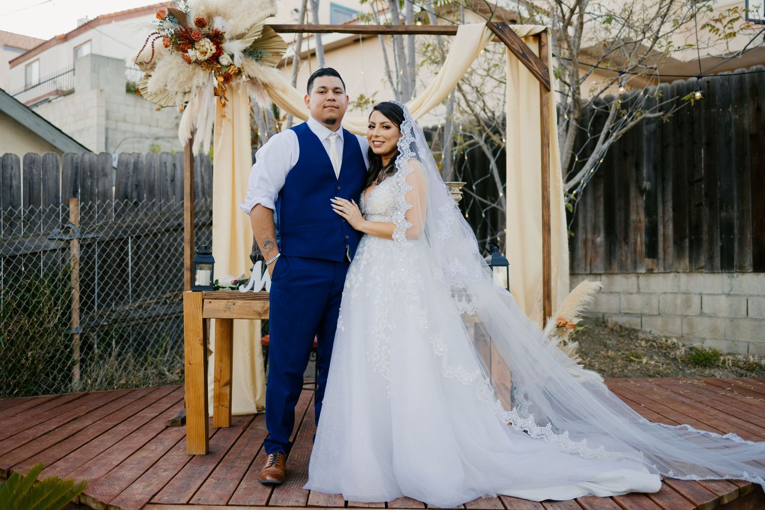 A wedding couple poses under a wooden arch decorated with floral arrangements.