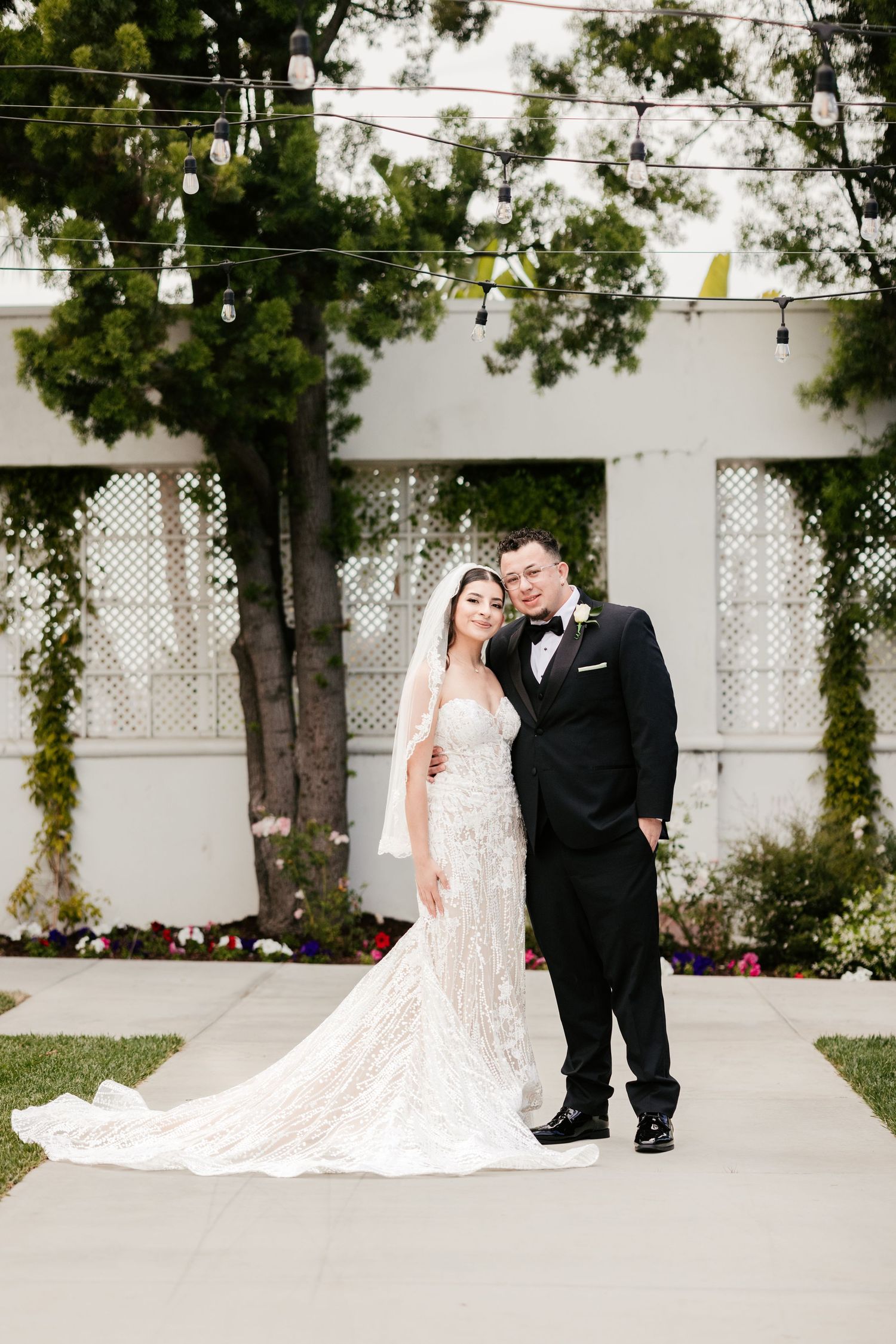 A romantic wedding portrait on a garden pathway with string lights and white lattice backdrop.