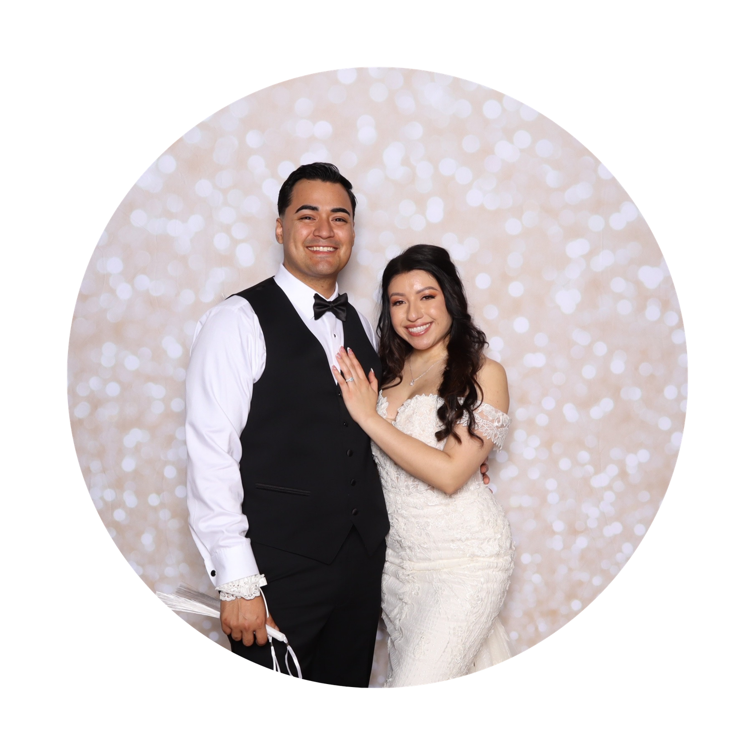 A couple poses in wedding attire, the groom in a black vest and the bride in a white lace gown, against a bokeh backdrop.