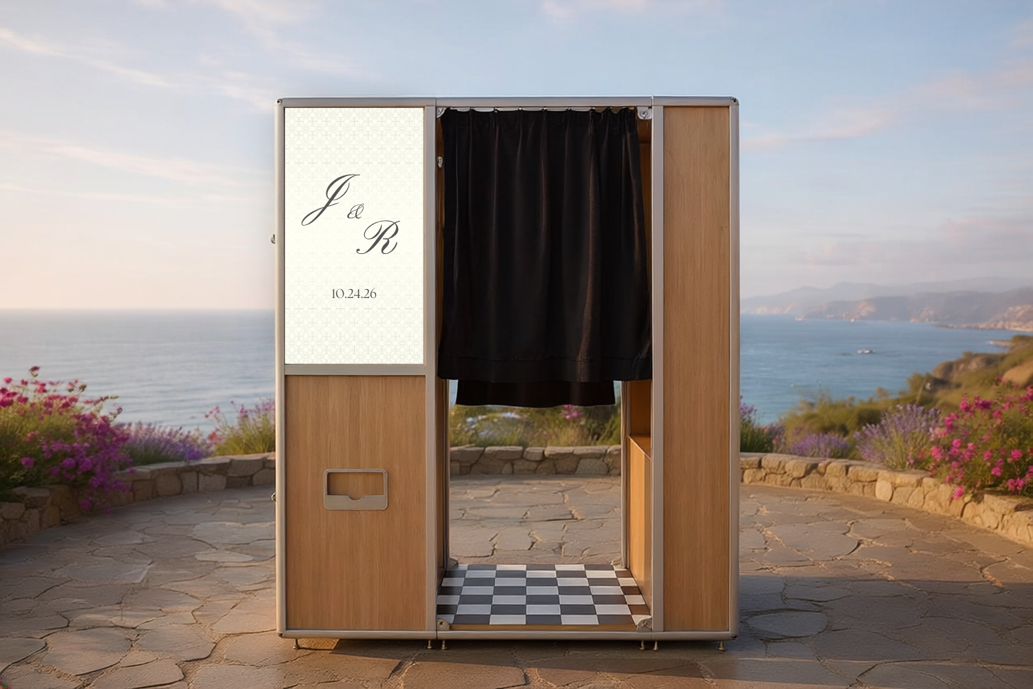 Elegant wooden photo booth with black curtain and monogram sign, set against a scenic ocean view at an outdoor event.