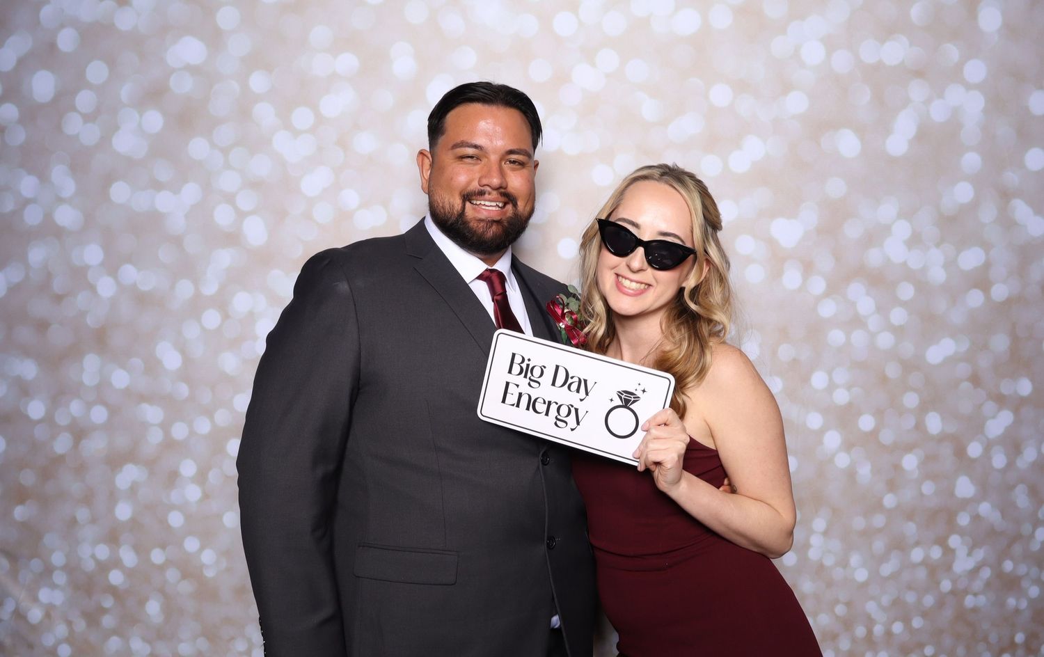 Couple posing at wedding photo booth, man in suit and woman in burgundy dress holding Big Day Energy sign.