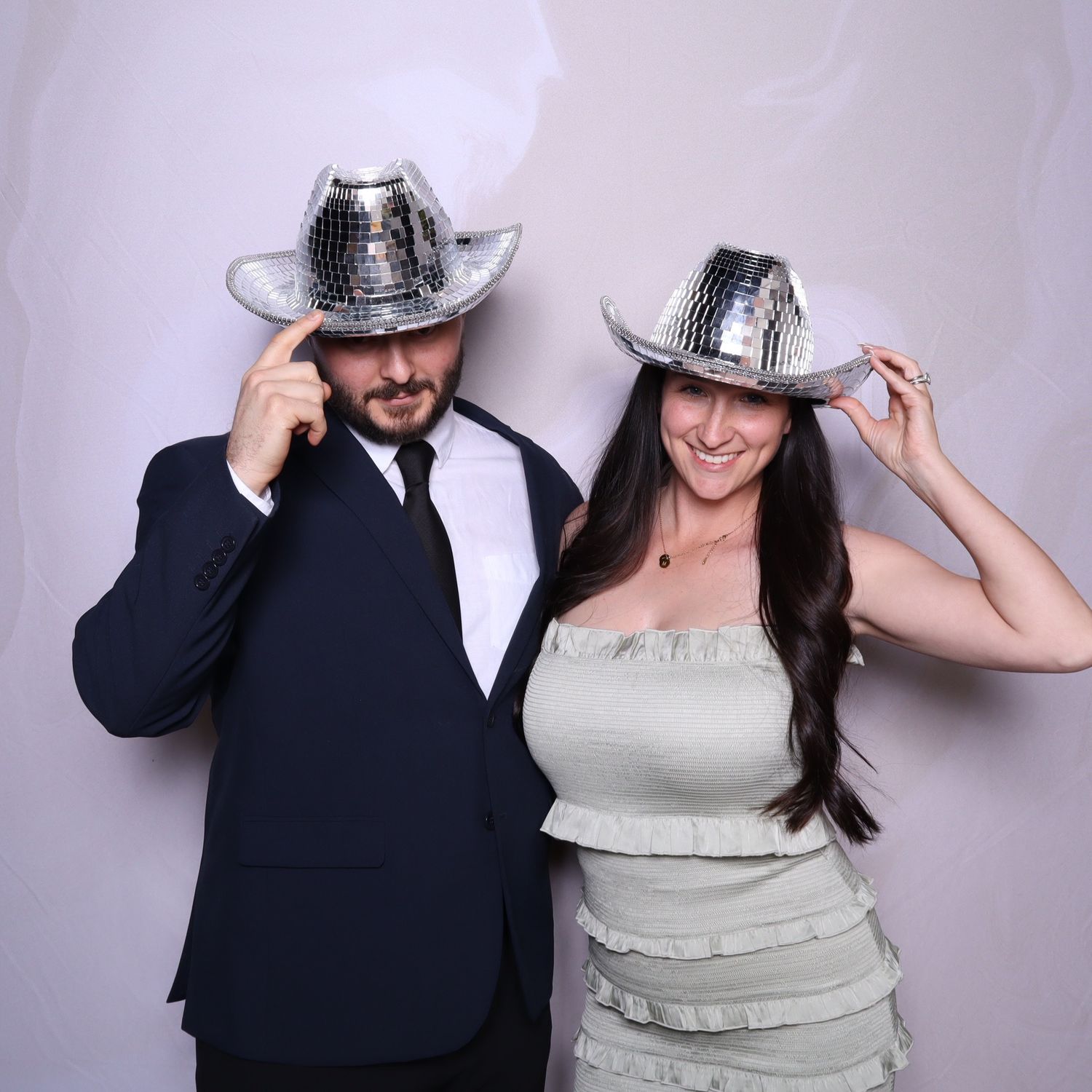 A man in a suit and a woman in a white dress pose together wearing silver sequin cowboy hats at a photo booth.