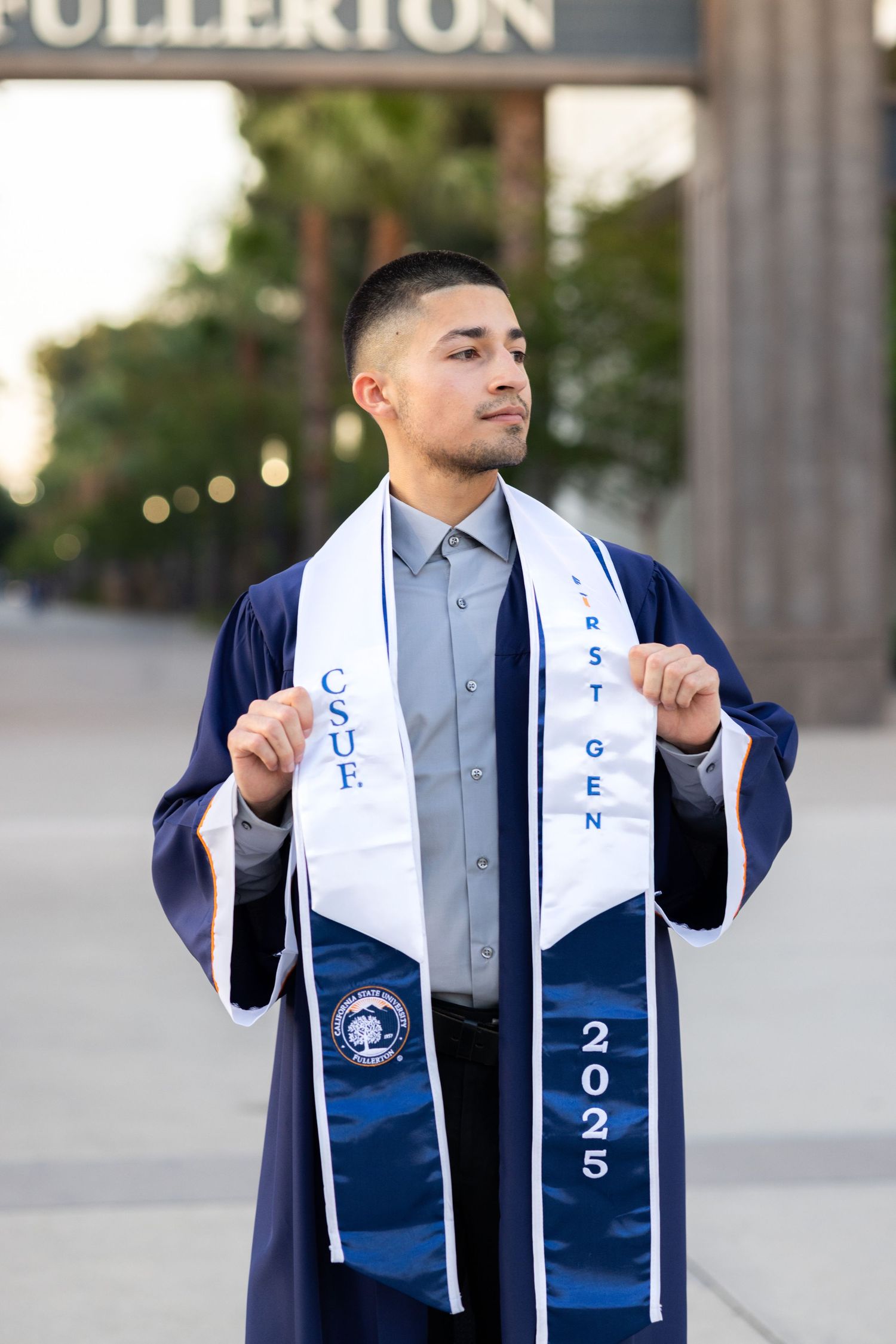 Graduate in navy blue regalia poses with graduation sashes at Fullerton campus.