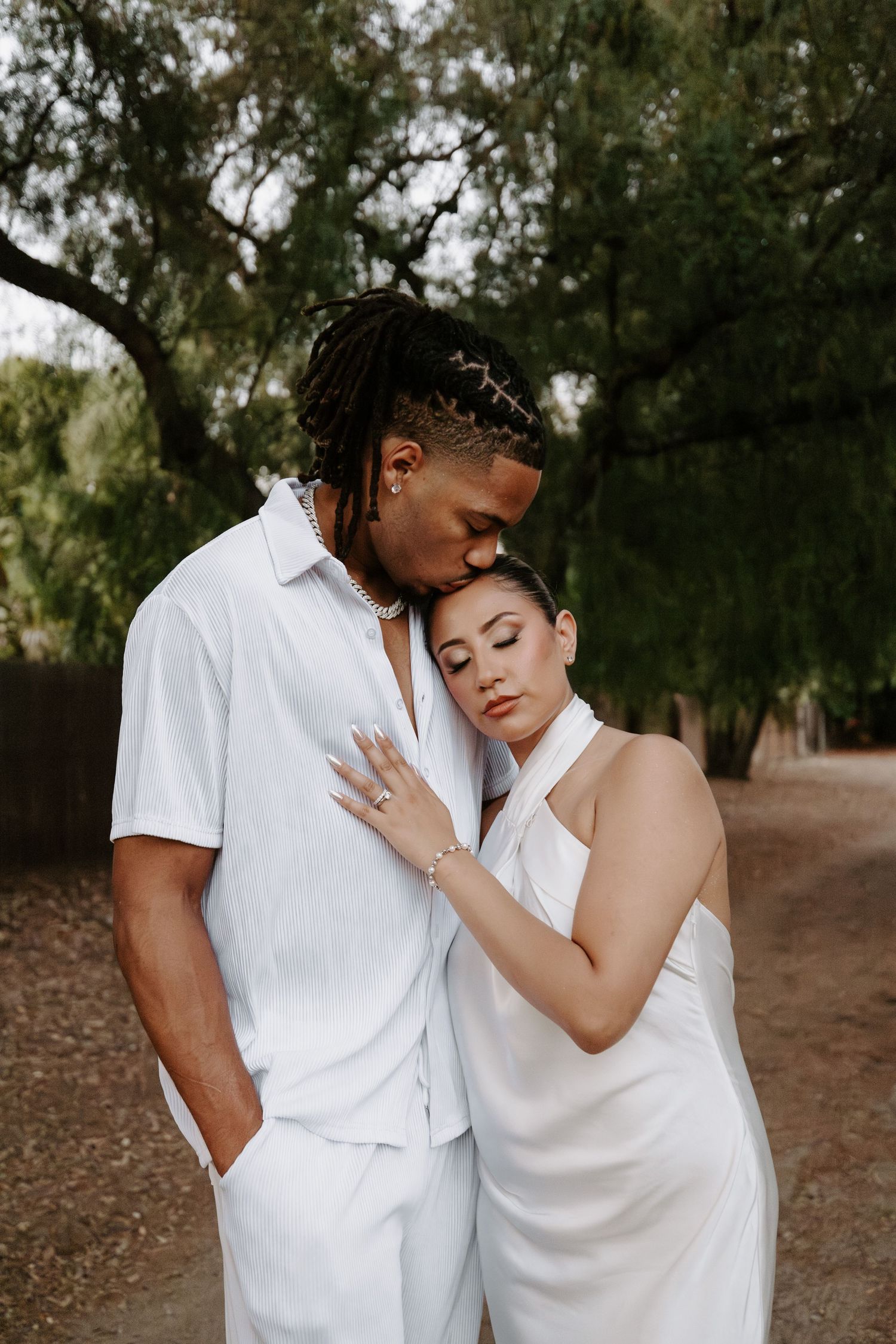 Intimate outdoor portrait of couple in all-white outfits embracing among trees.