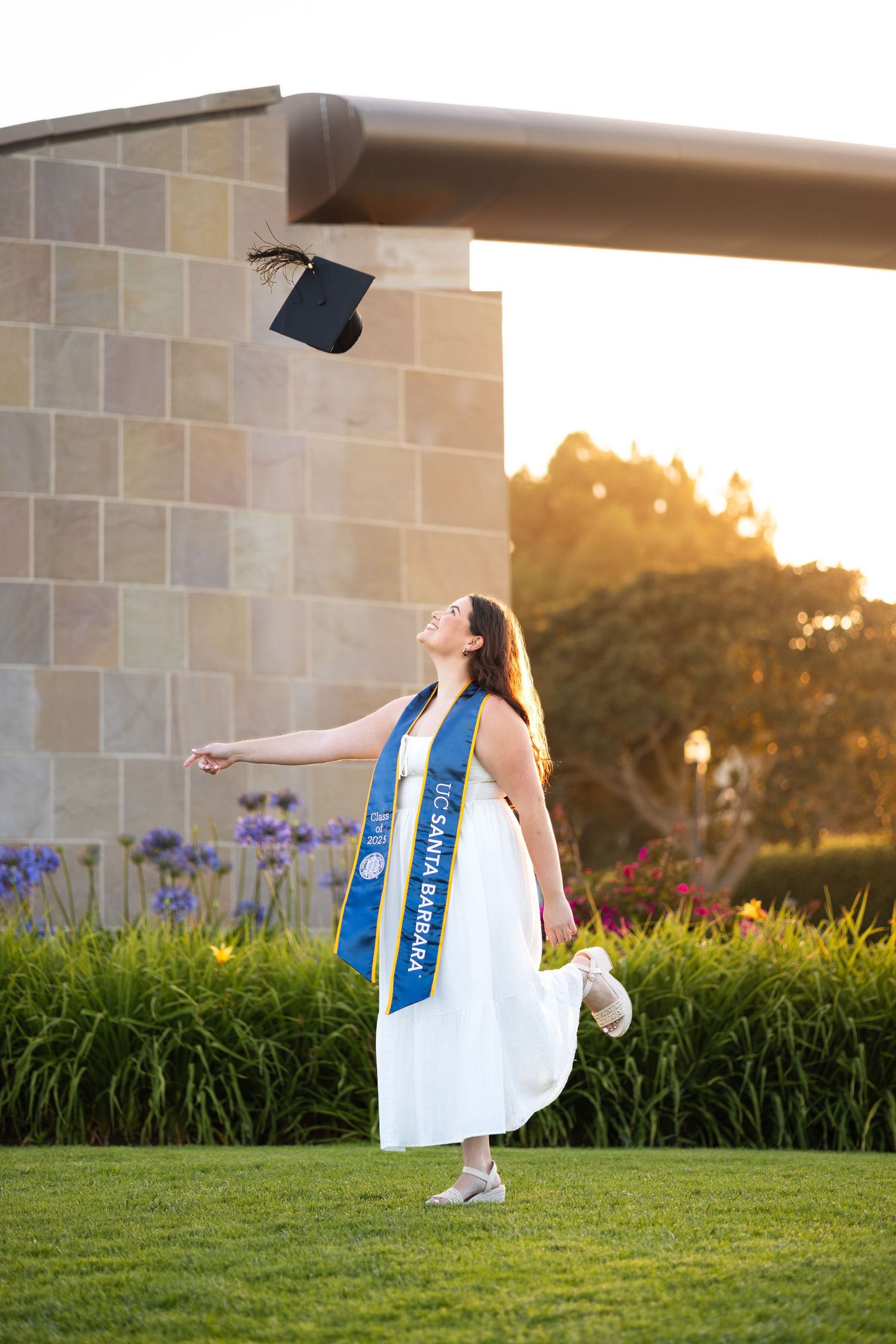 Graduate in white dress and blue sash poses on grass during golden hour celebrations.