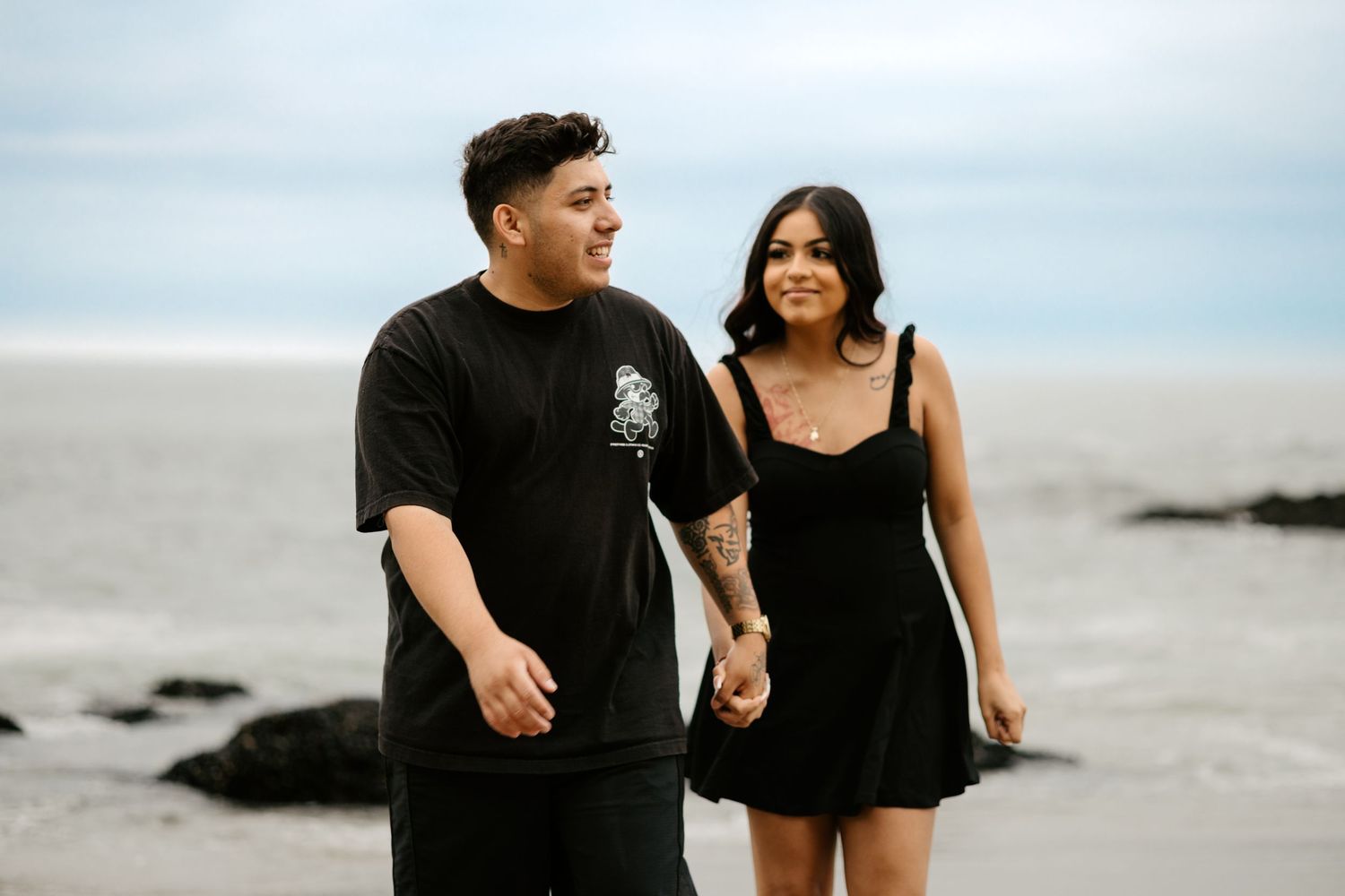 Couple walking hand in hand on rocky beach wearing black outfits against cloudy sky.