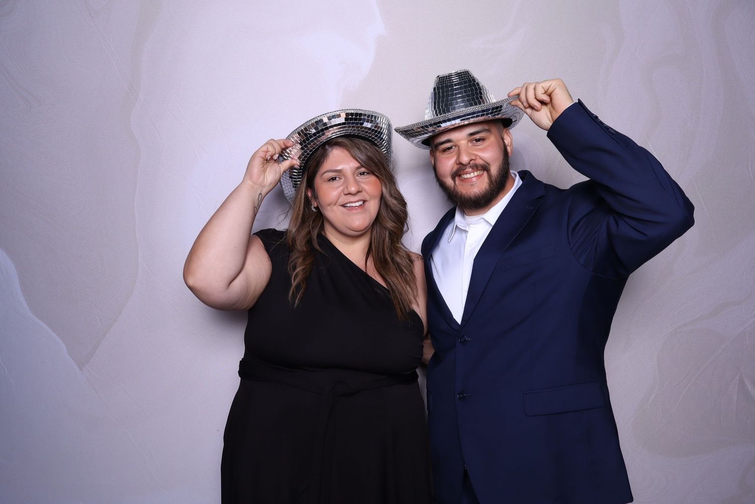 A woman in black dress and man in navy suit pose playfully with novelty hats at a photo booth event.