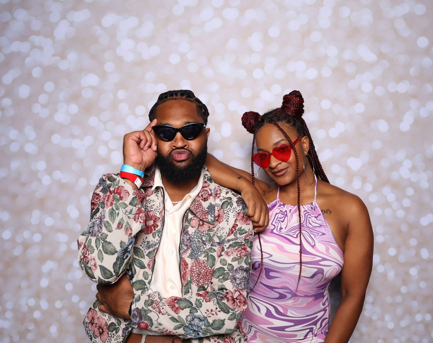 A man in a floral shirt and a woman in a purple dress pose together wearing red sunglasses at a photo booth.