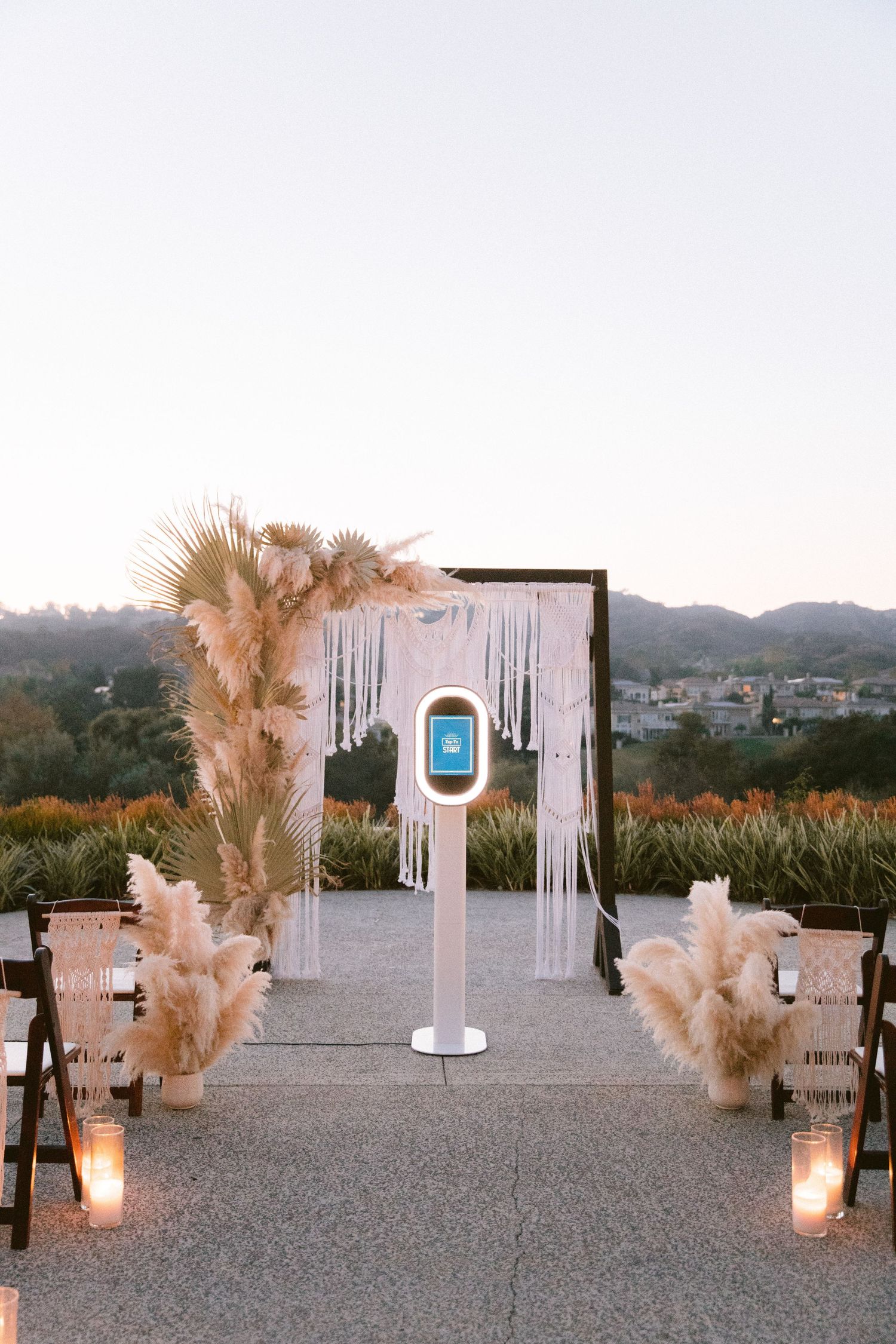 Outdoor wedding ceremony setup with white panels, pampas grass arrangements, and glowing candle lanterns at sunset.