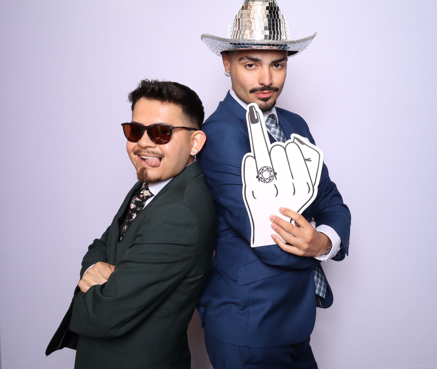 Two men in suits pose back-to-back, one in sunglasses, the other in a disco cowboy hat holding a foam finger.