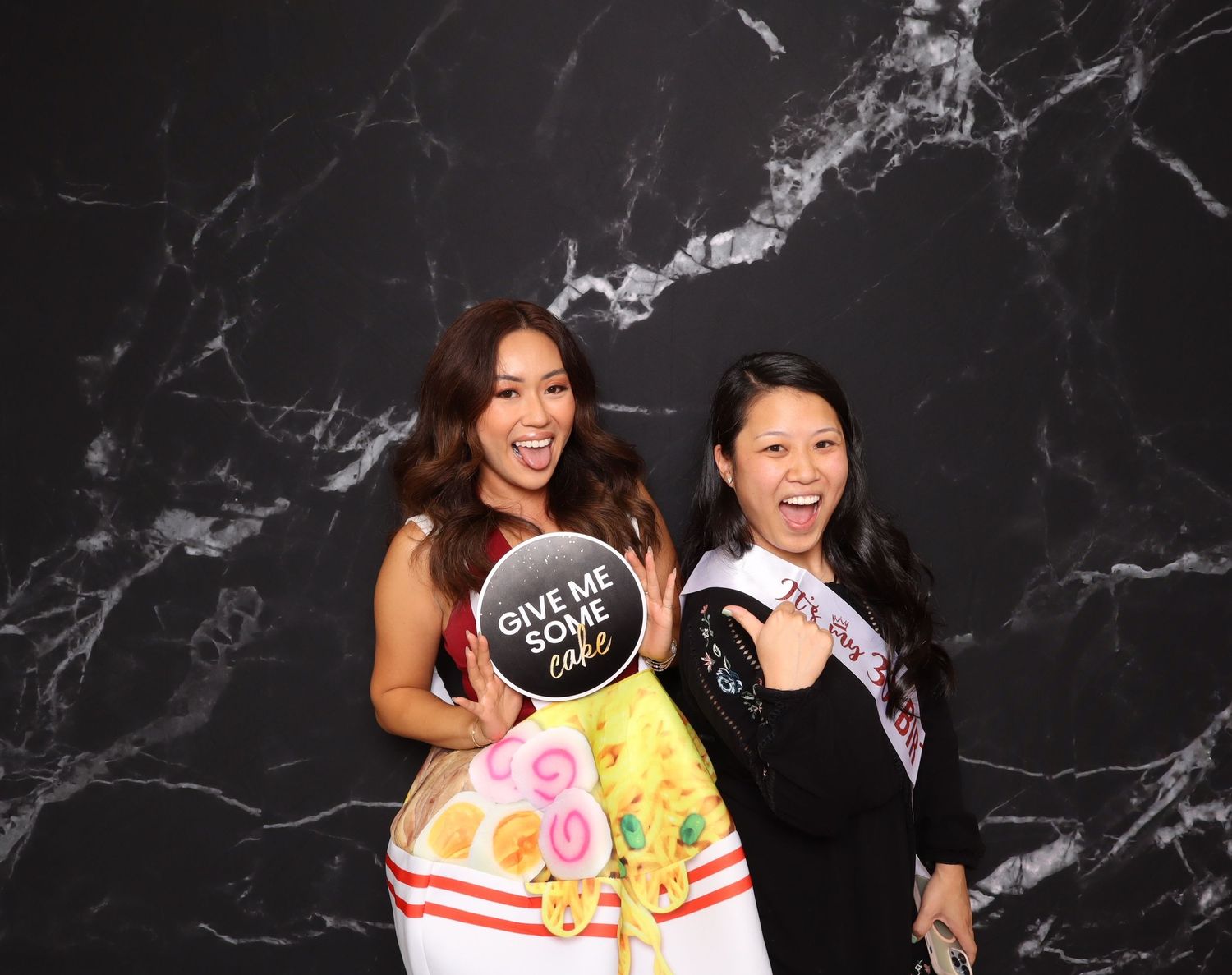Two women posing and smiling at a photo booth with marble backdrop, one holding a 'Give Me Some Cake' sign.