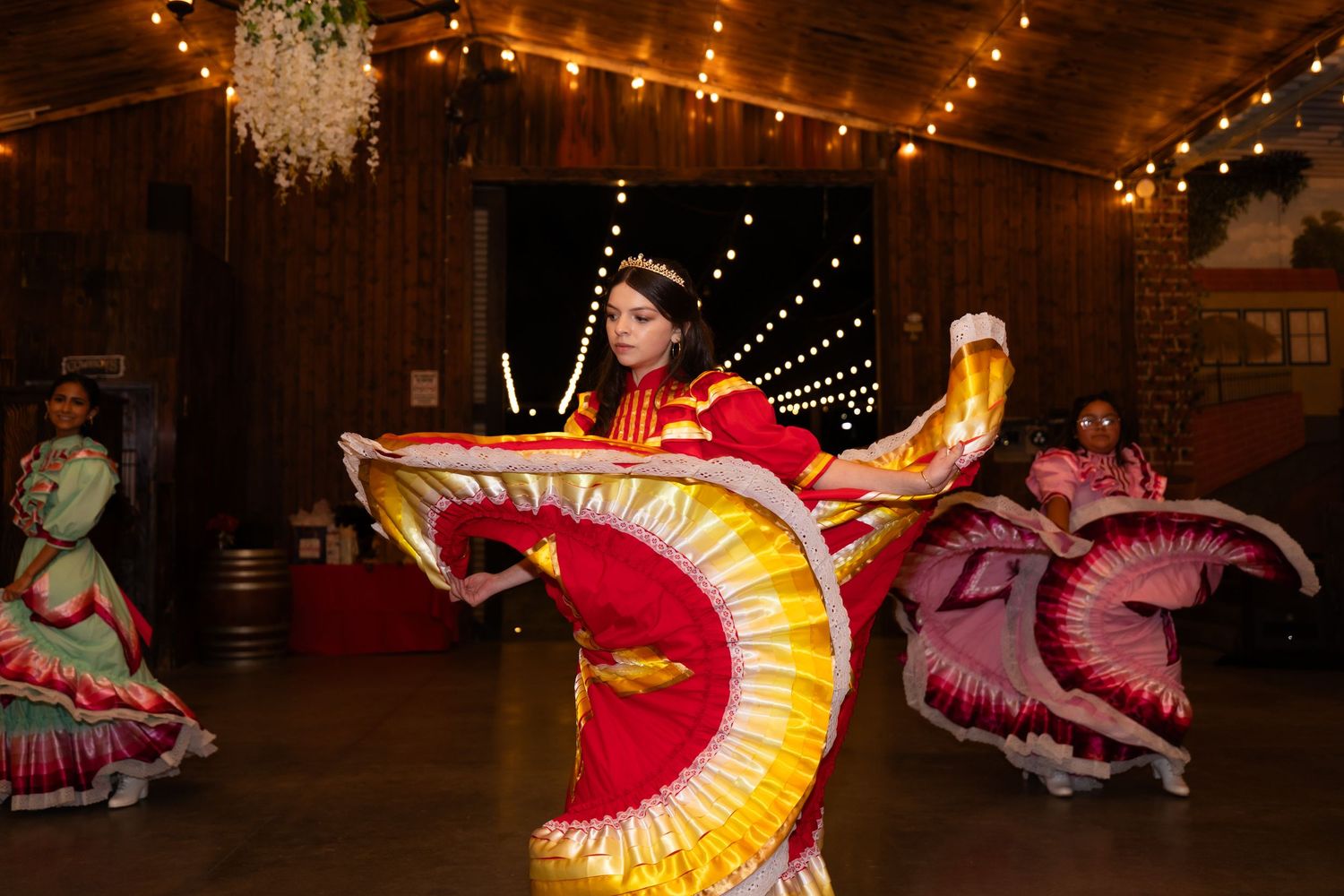 Traditional dancers in vibrant spinning dresses performing under indoor string lights.