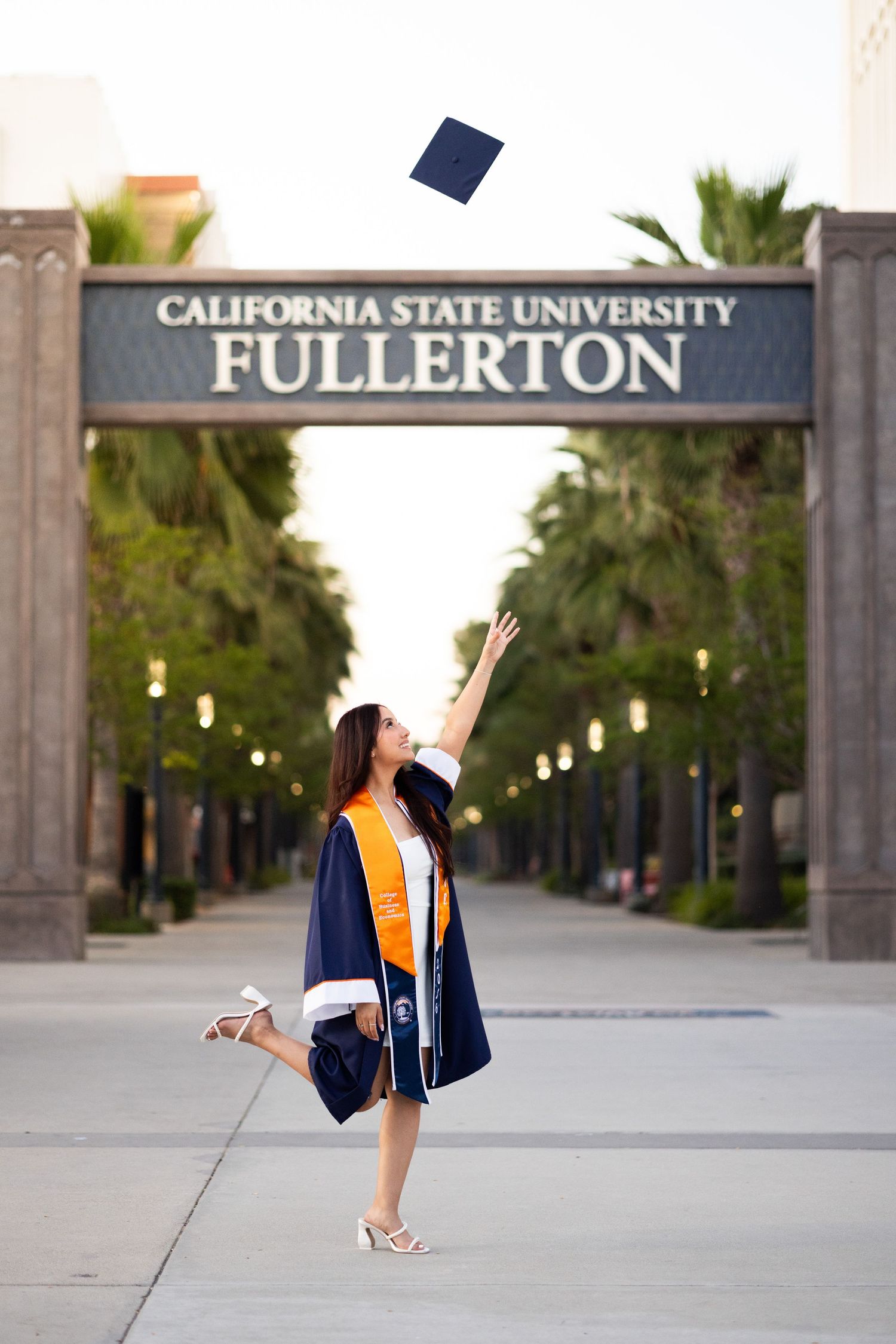 Graduate in navy cap and gown tosses mortarboard at California State University Fullerton entrance arch.