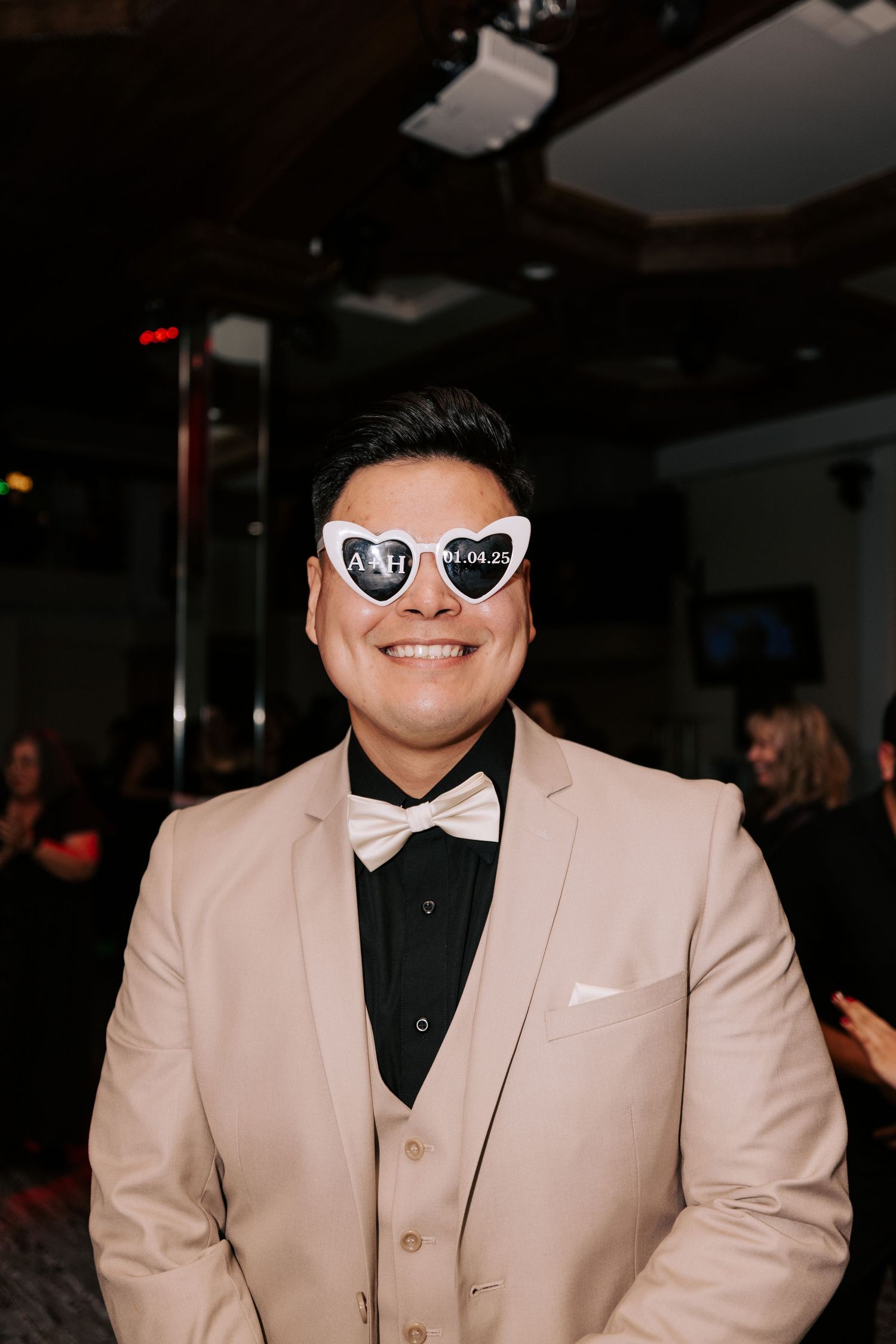 A person in a beige tuxedo and bow tie wearing heart-shaped sunglasses at a celebration.
