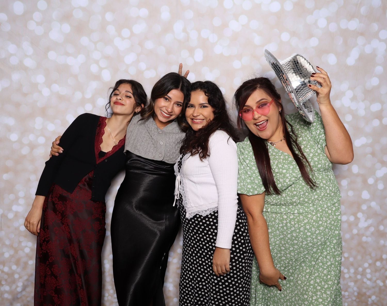 Four women posing together at a photo booth, smiling, one holding a disco ball hat at a festive party event.