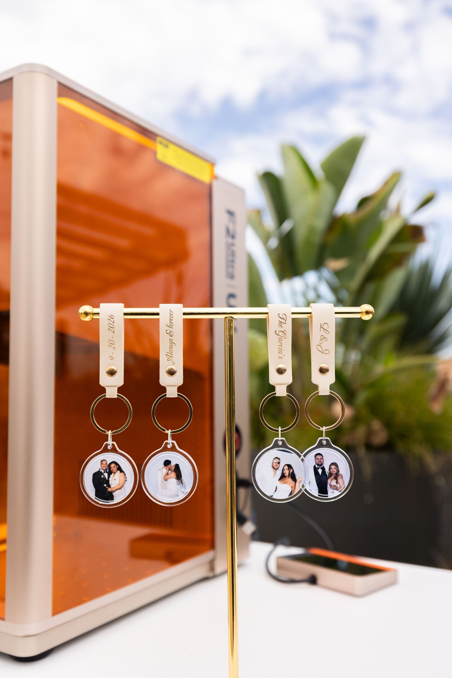 Custom photo keychains with couple portraits on a gold display stand, next to an orange laser engraver outdoors.