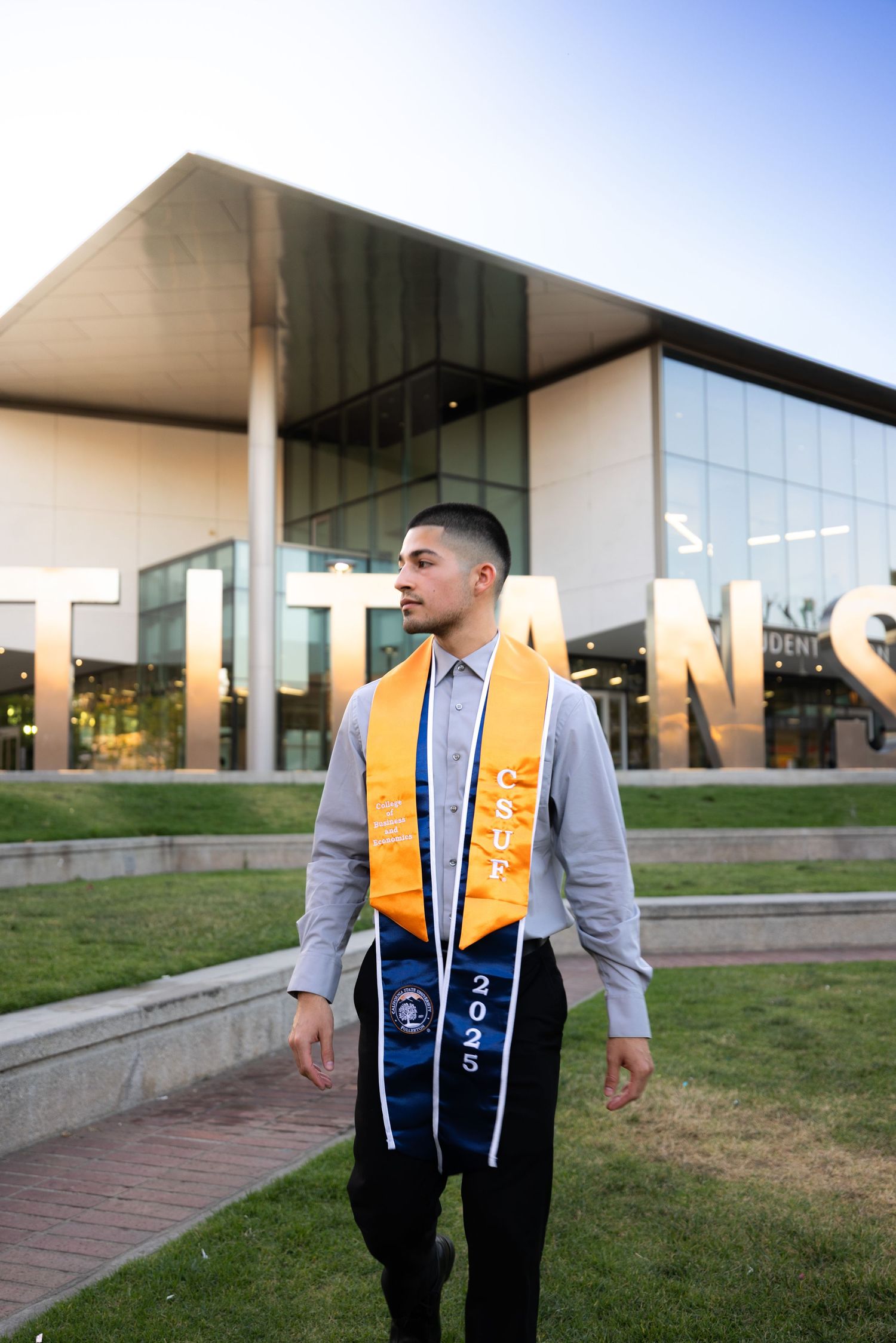 Graduate in grey shirt and graduation stole stands on campus steps during golden hour.